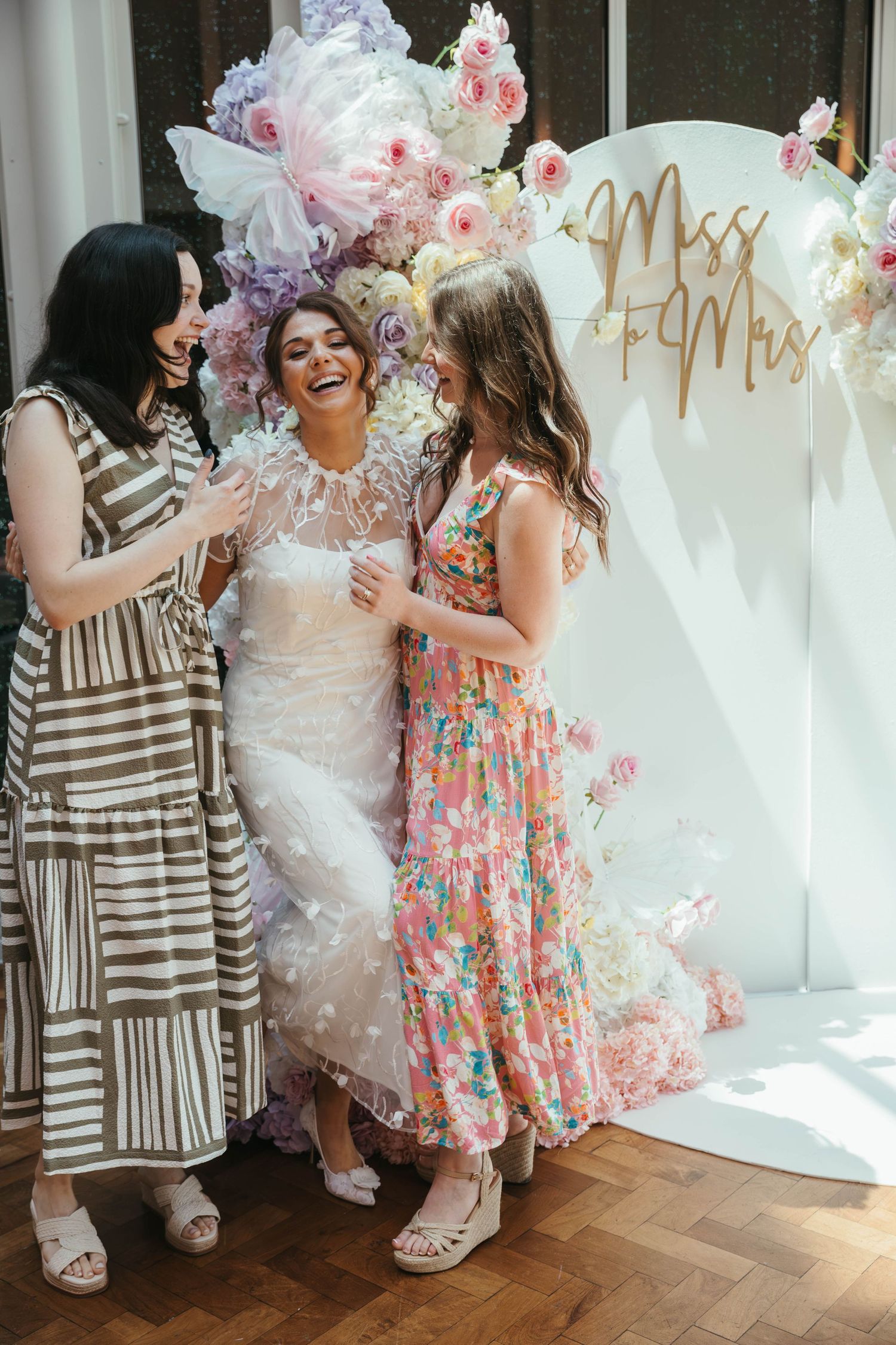 Bride in white lace gown laughing with two friends at a Miss to Mrs bridal shower photo backdrop.