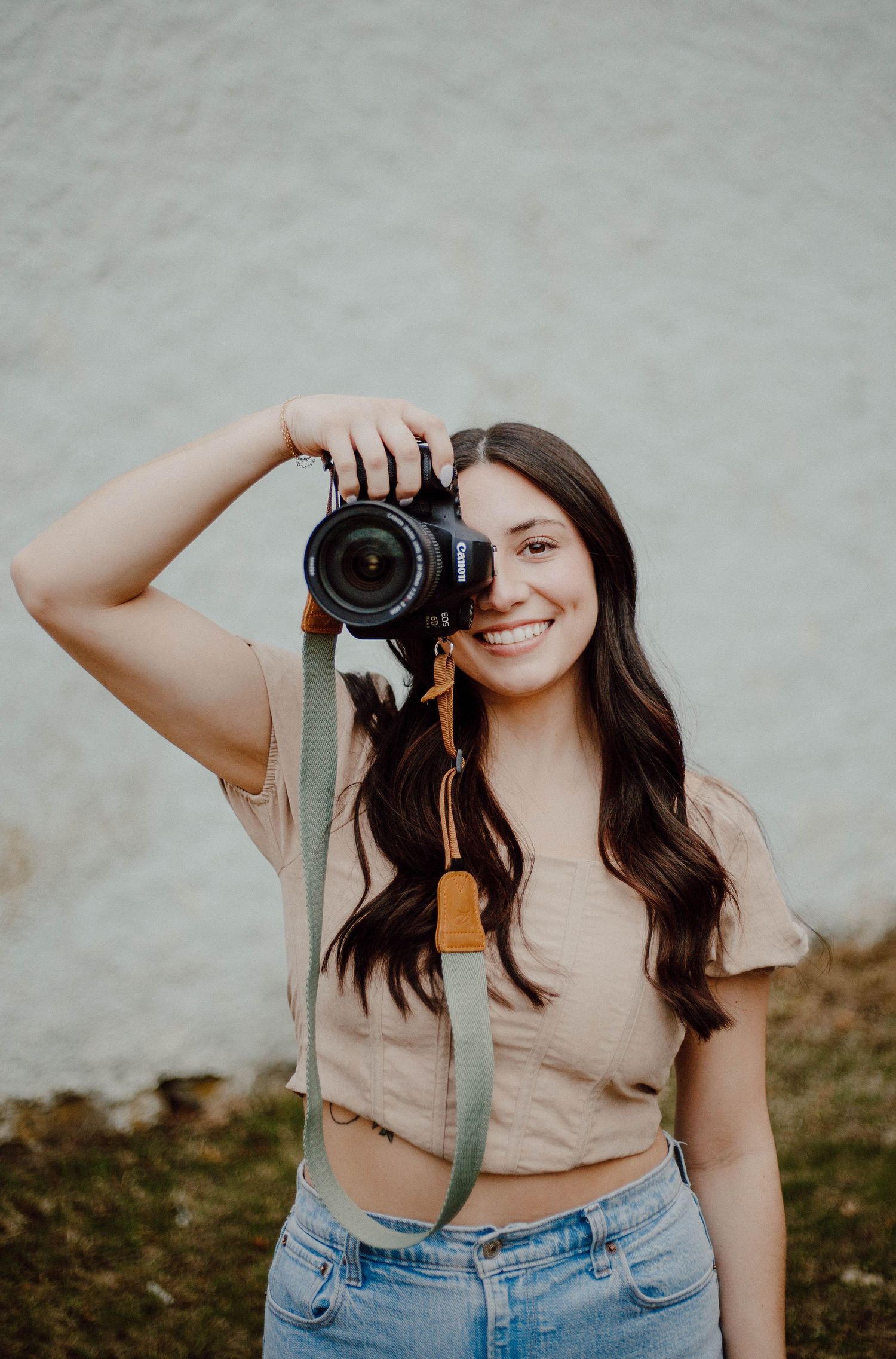 Smiling young woman with dark hair holding DSLR camera up to face with green strap outdoors.