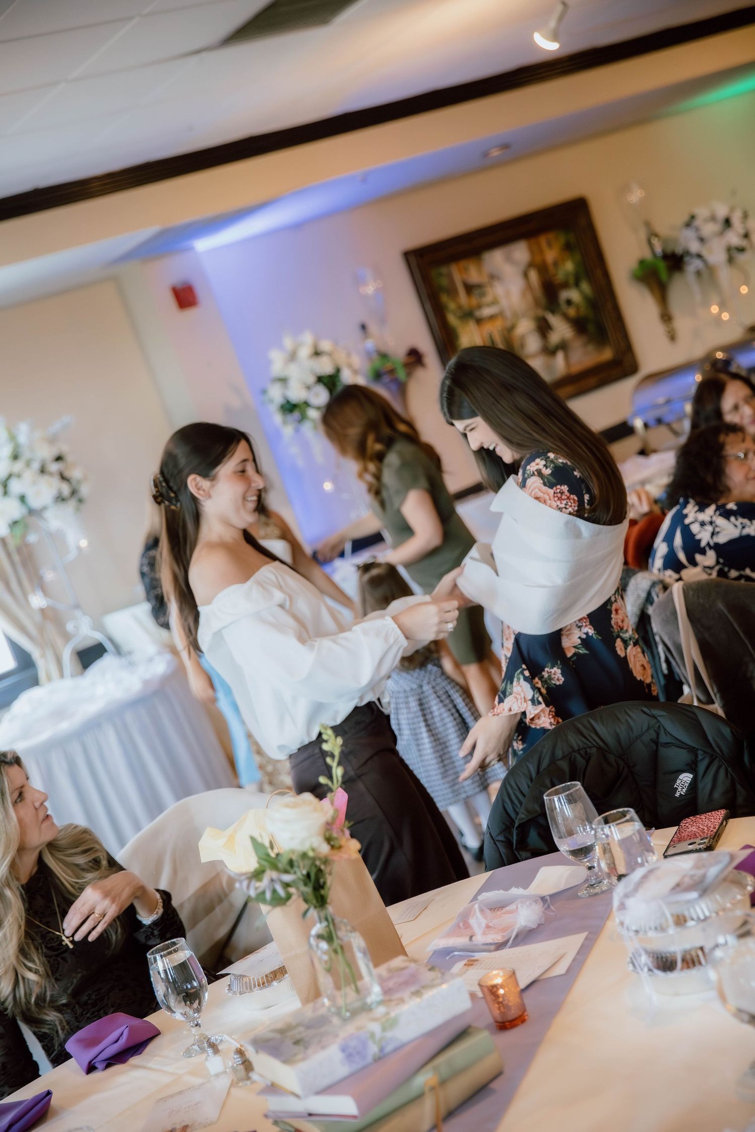 Two women sharing a moment at an elegant wedding reception with floral centerpieces and purple accents.