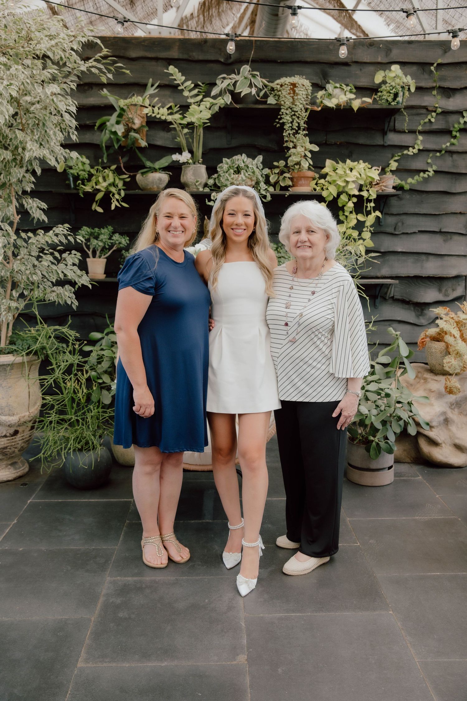 Three women smiling together outdoors in front of a lush green plant wall on a stone patio.