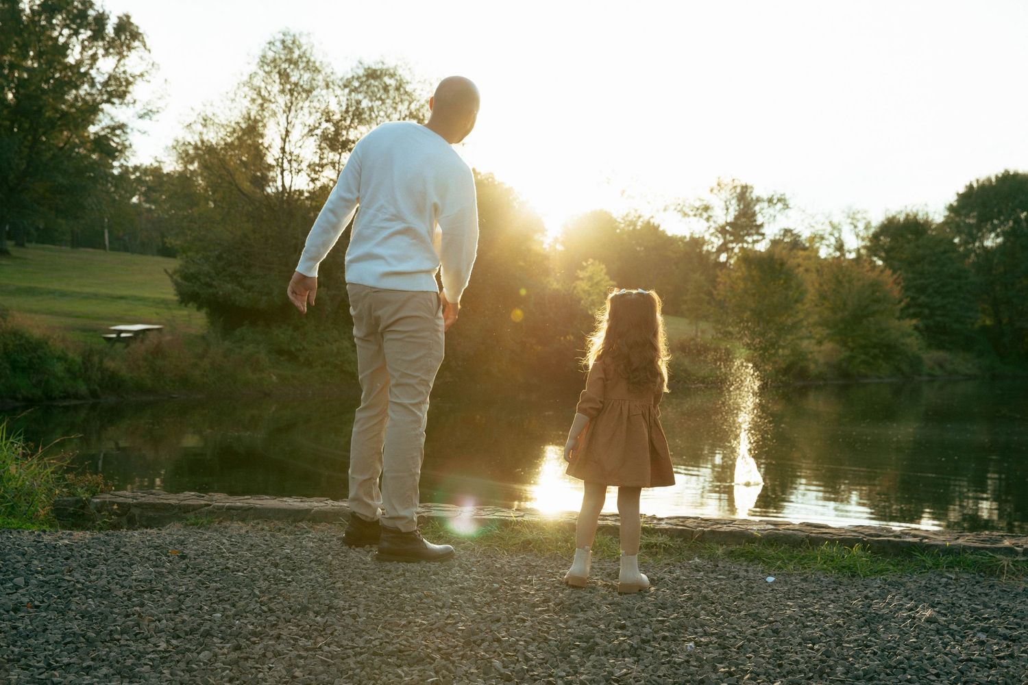 Adult and child holding hands by a sunlit lake at golden hour, surrounded by lush green trees.