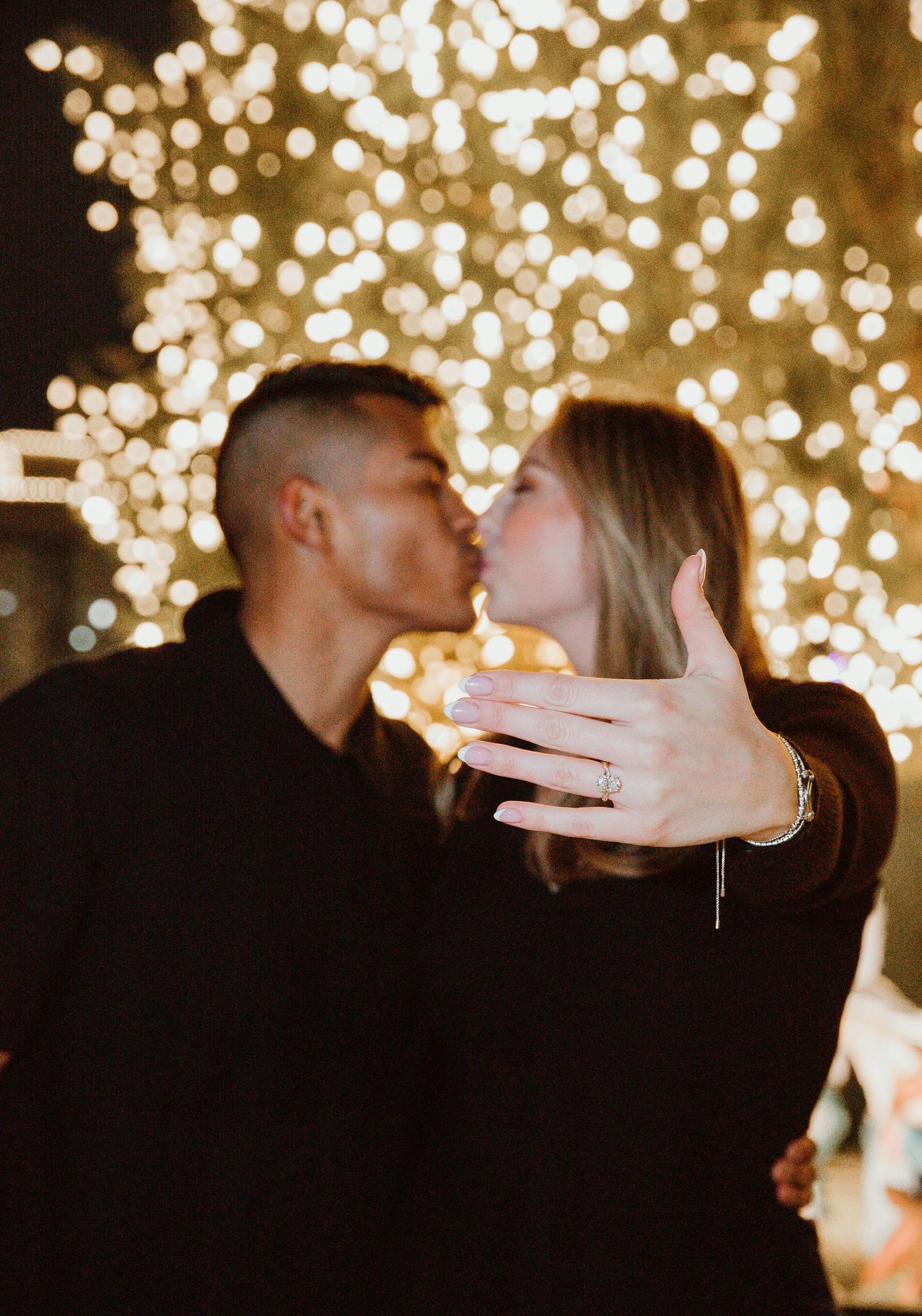 Couple kissing in front of bokeh Christmas lights, woman showing off engagement ring to camera.