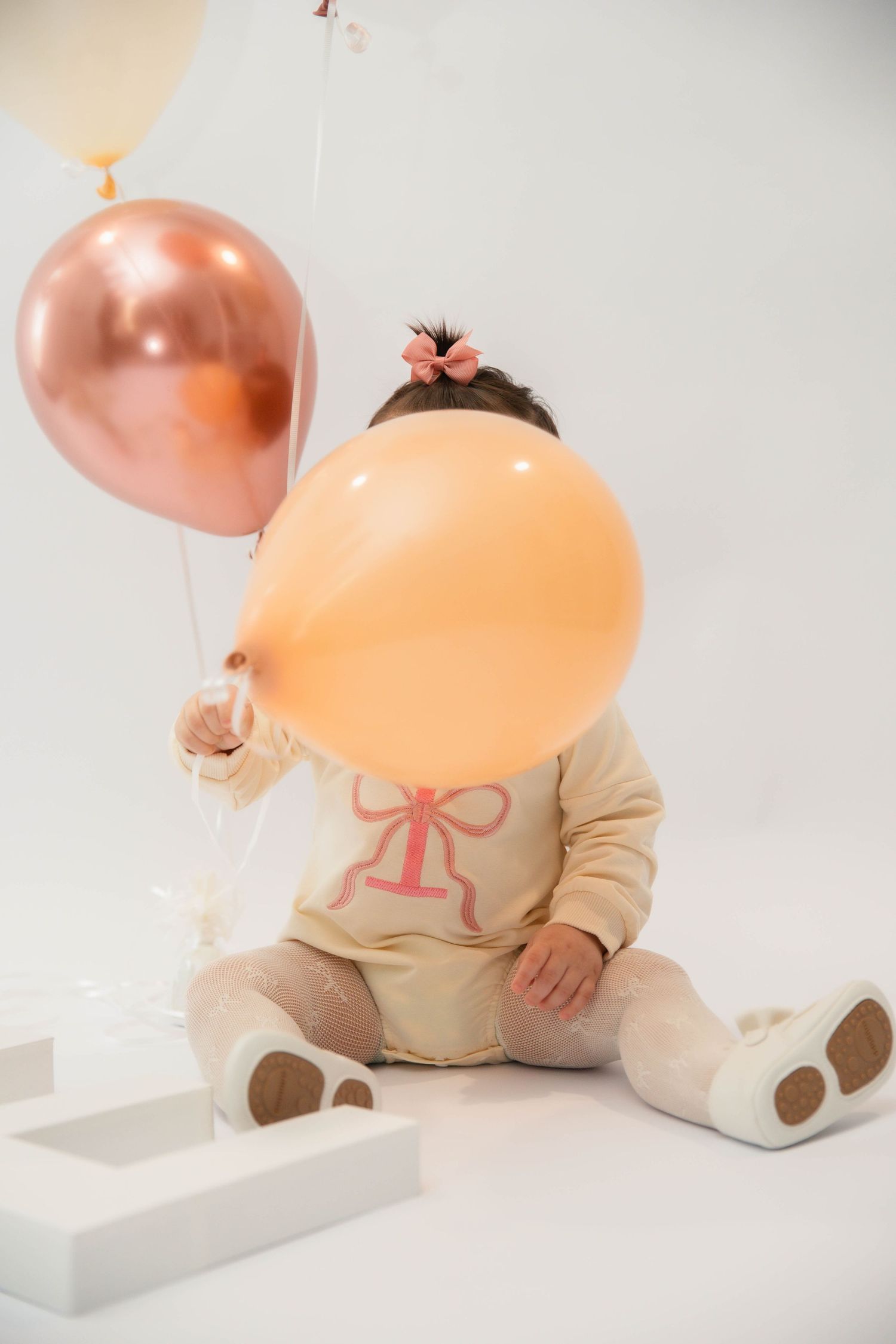 Baby girl in cream outfit sitting with peach and rose gold balloons, wearing bear booties, celebrating first birthday.