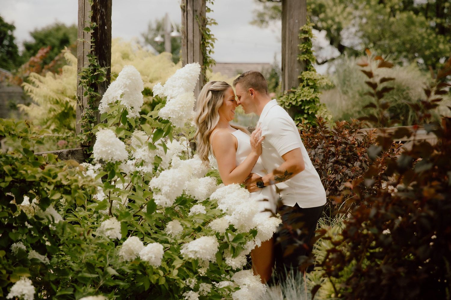 Couple sharing a kiss surrounded by lush white hydrangea blooms in a garden setting.