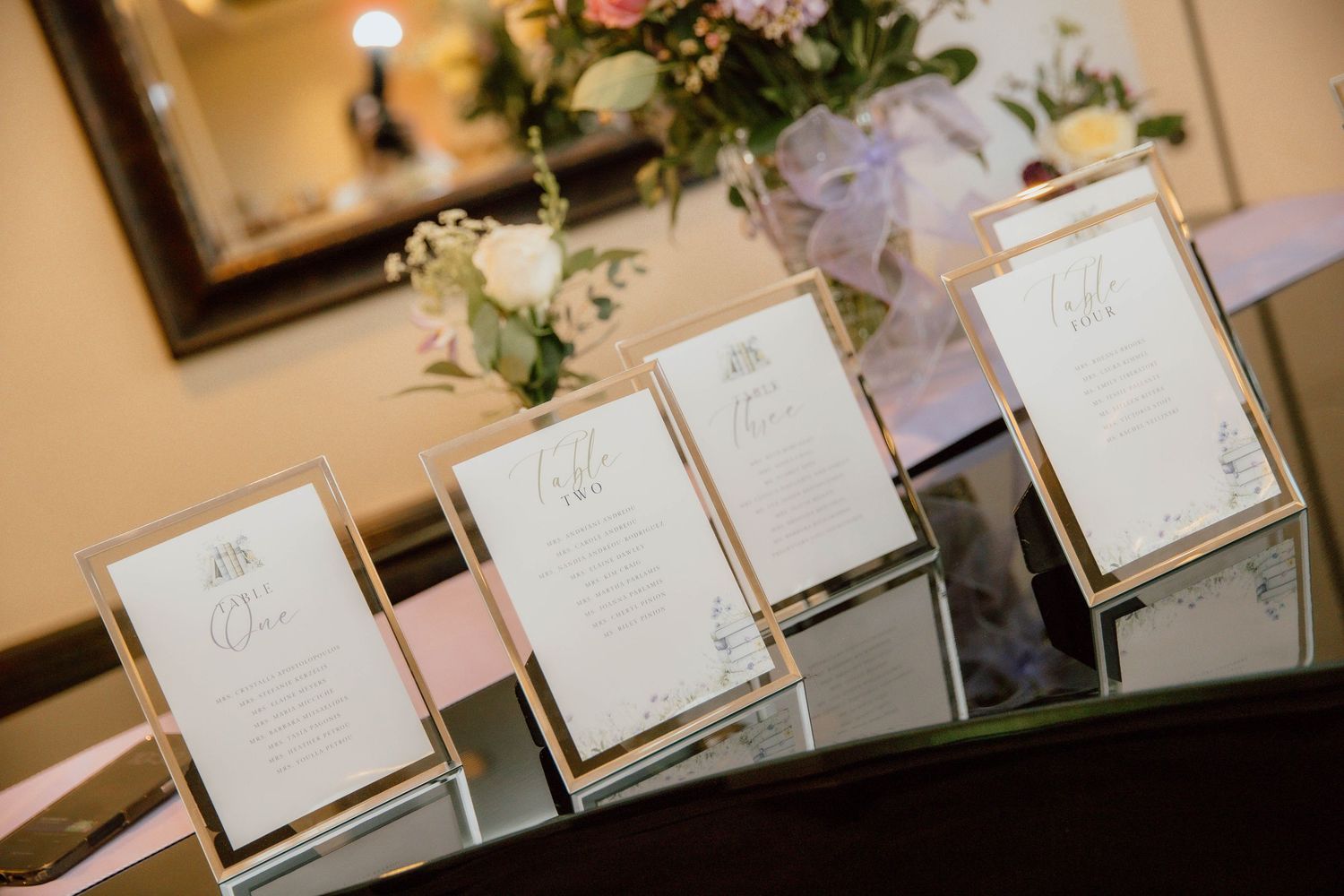 Gold-framed wedding table number cards displayed on a mirrored surface with floral arrangements at an elegant reception.