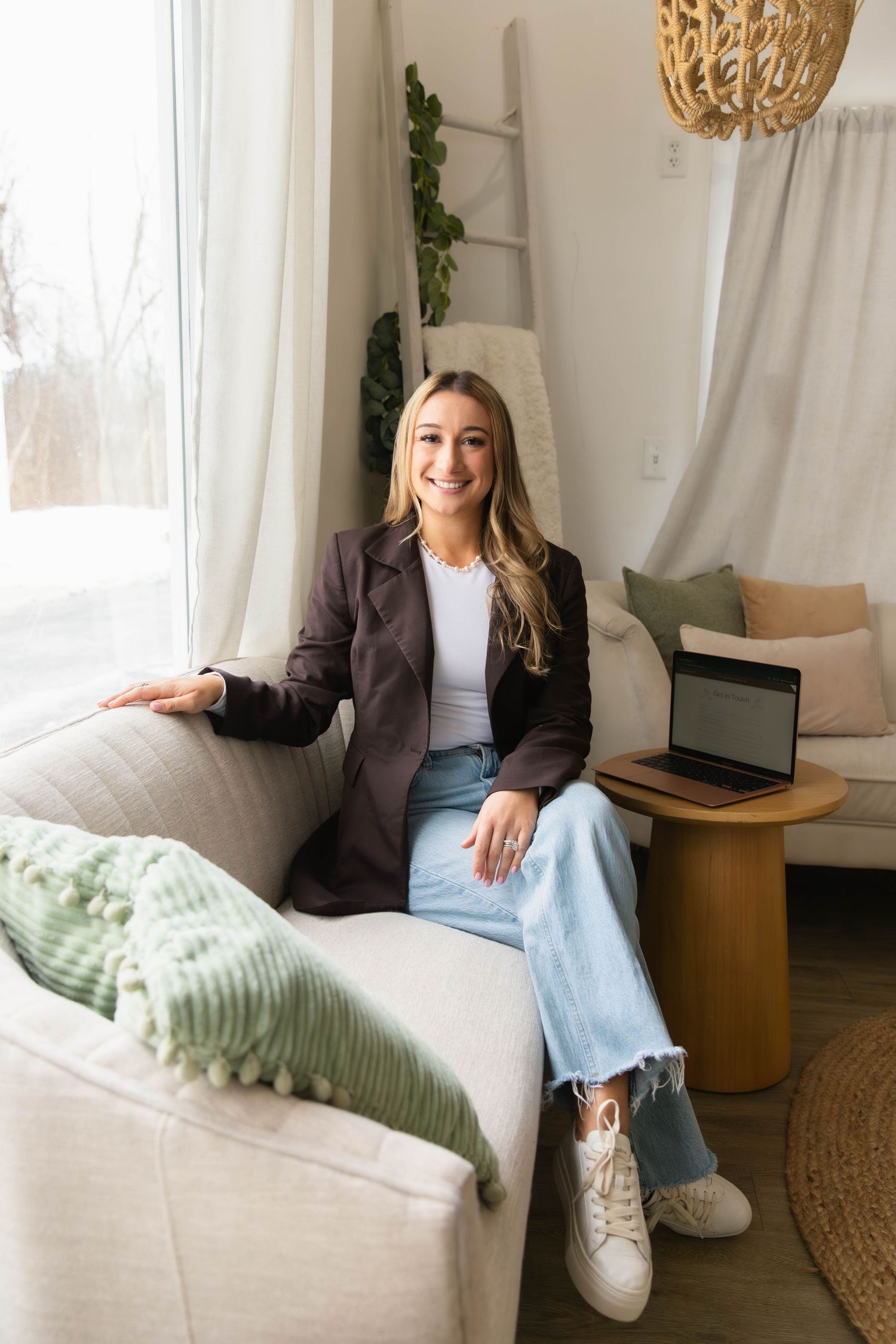 Smiling blonde woman in blazer and jeans sits on a couch beside a laptop in a modern living room.