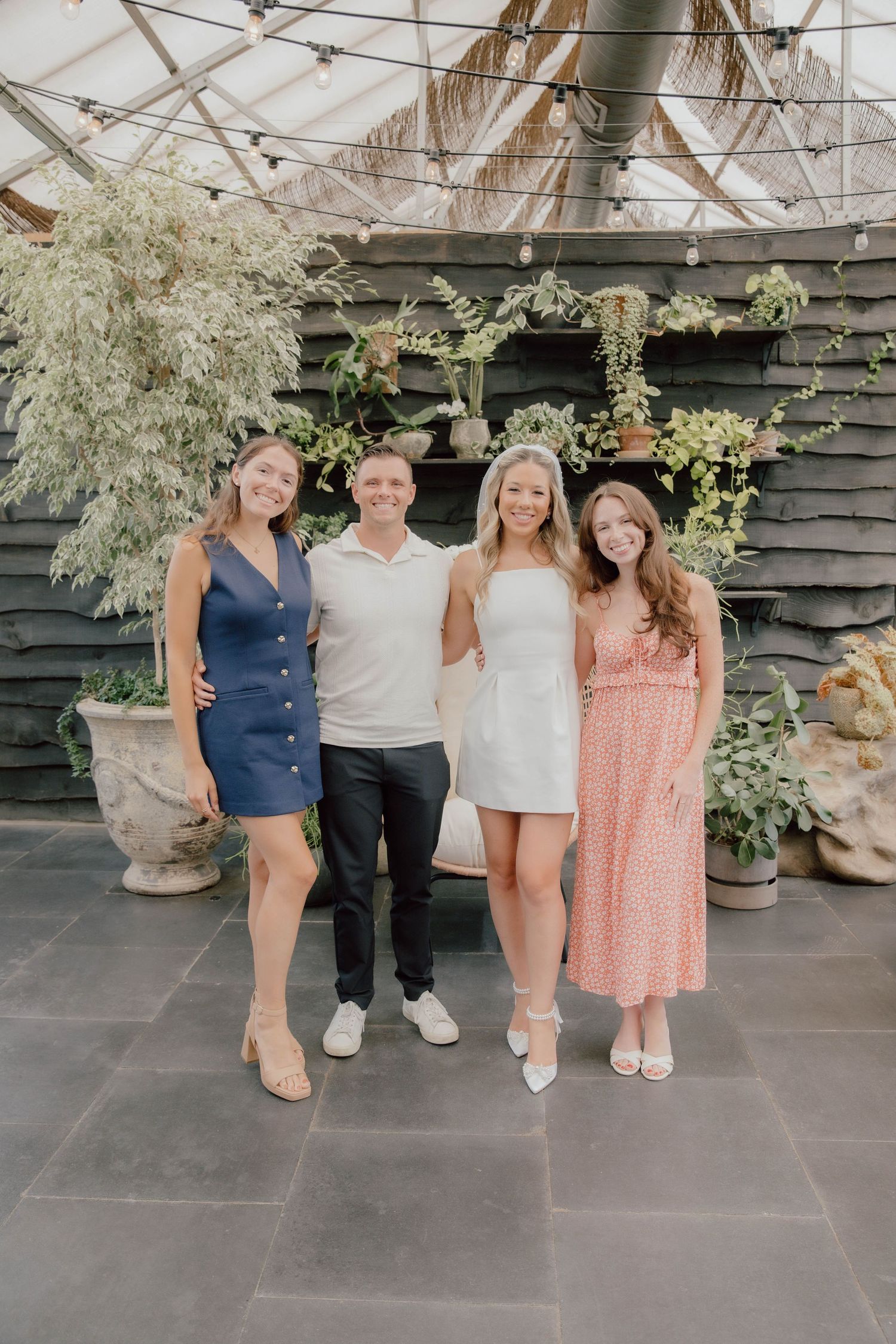 Four people posing together in a greenhouse venue decorated with hanging plants and string lights.