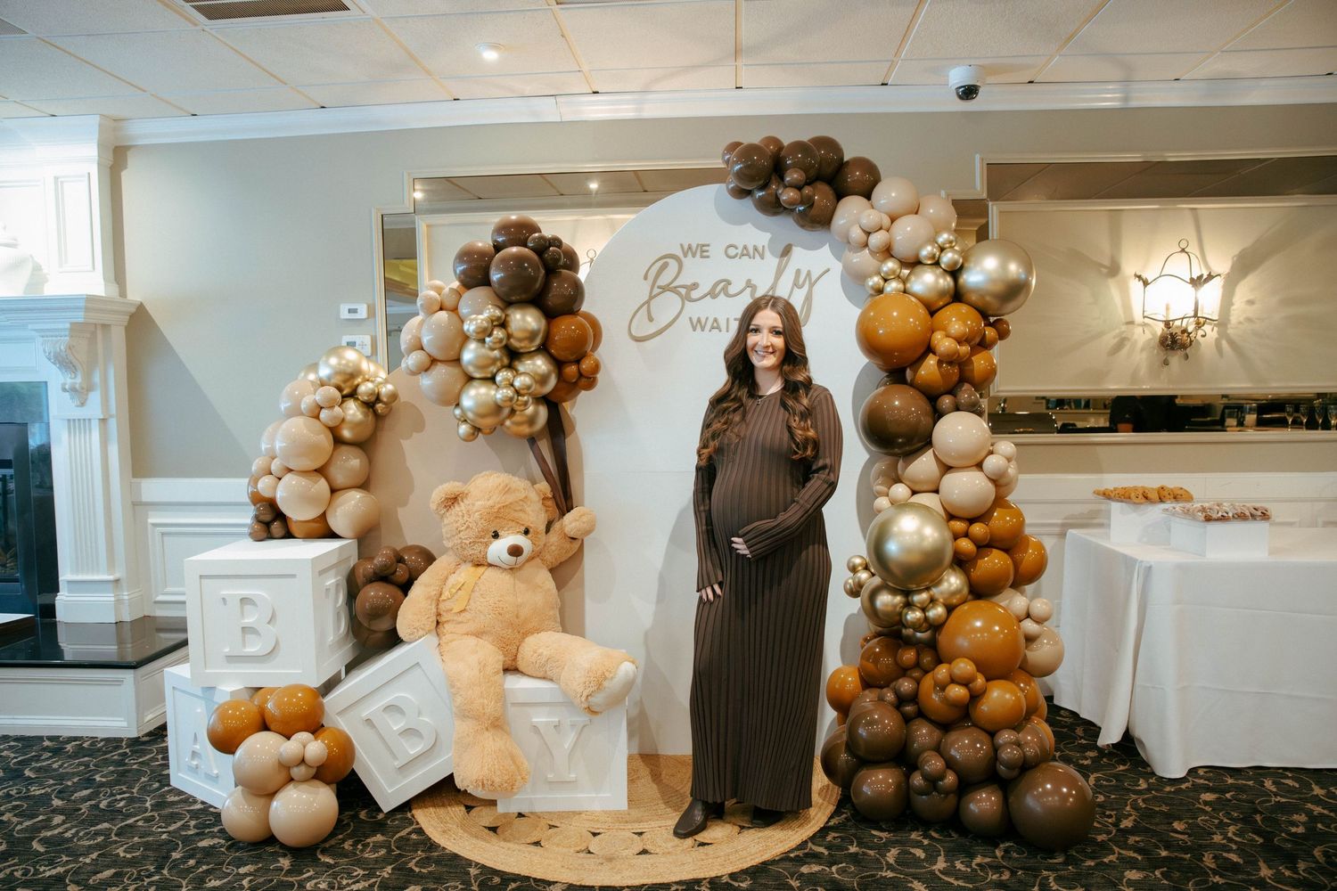 Pregnant woman posing at baby shower beside giant teddy bear and brown balloon arch display.