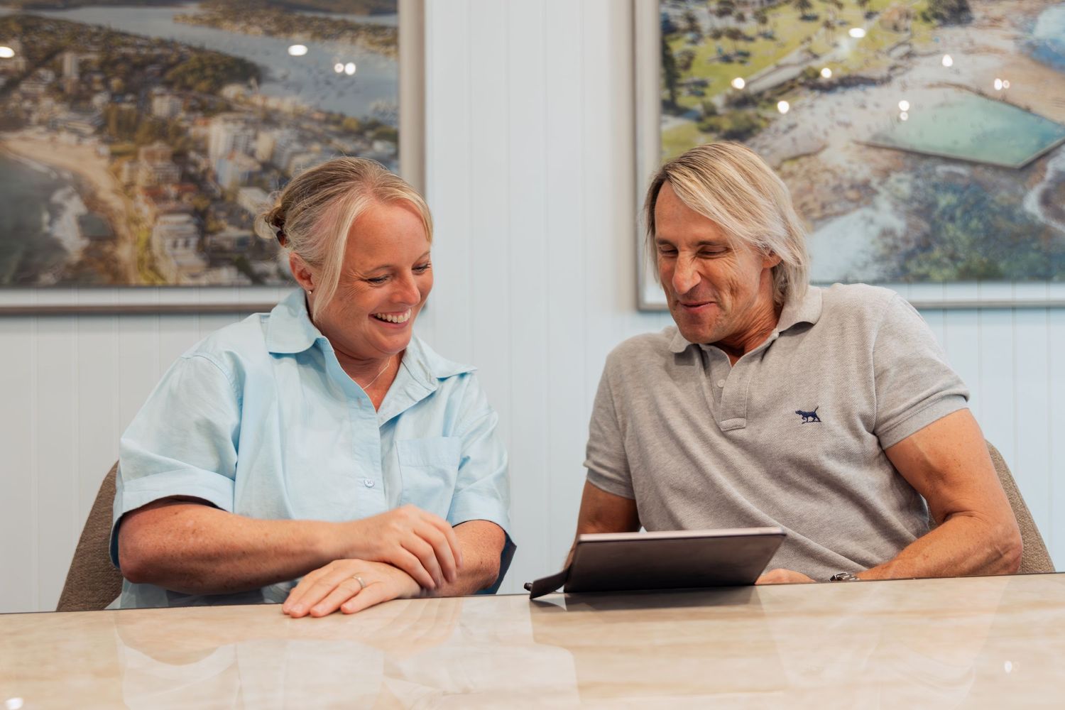 Two professionals smiling while reviewing content on a tablet at a modern office table with coastal artwork in background.
