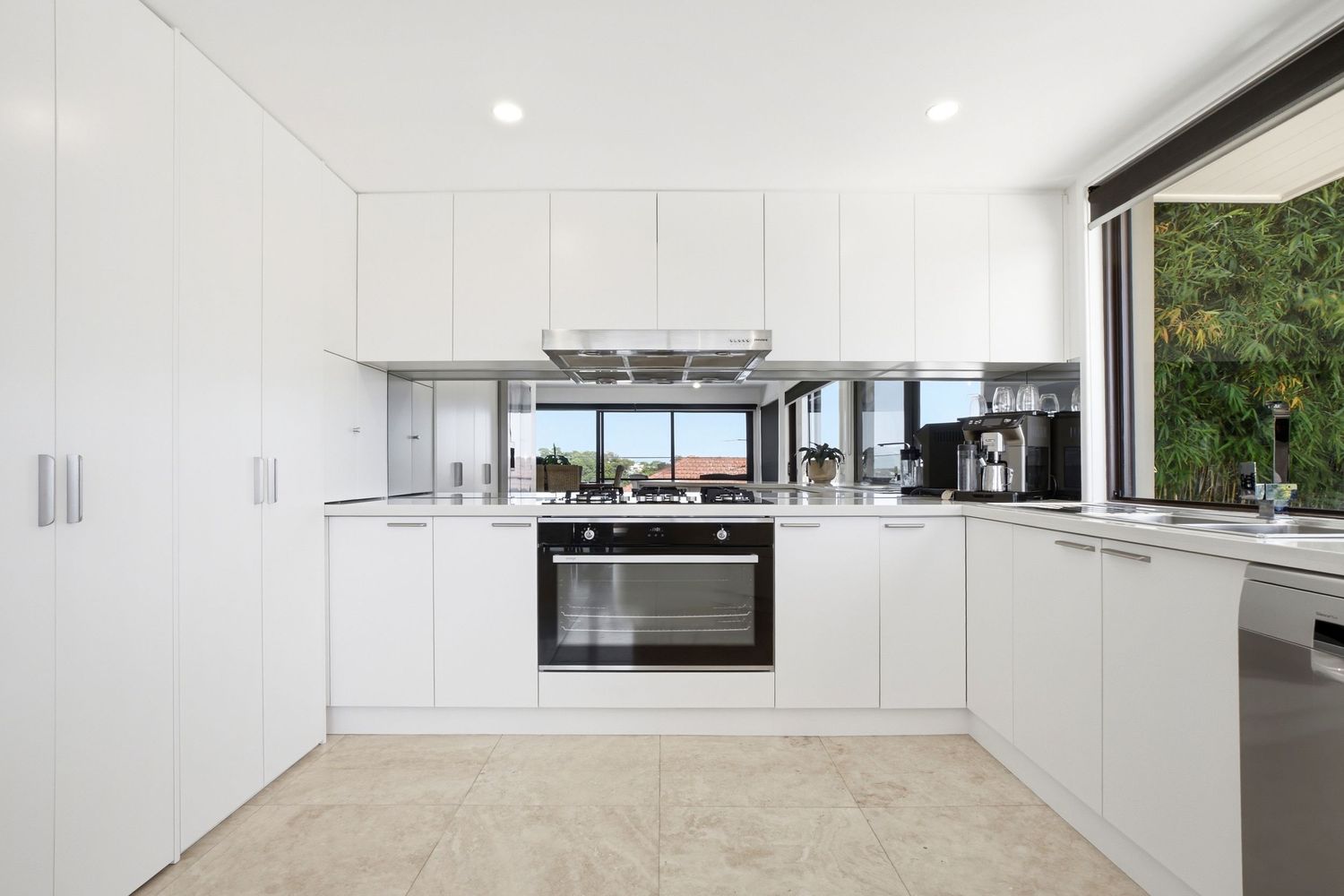 Modern white kitchen with built-in oven, mirrored splashback, sleek cabinetry, and large window with garden view.