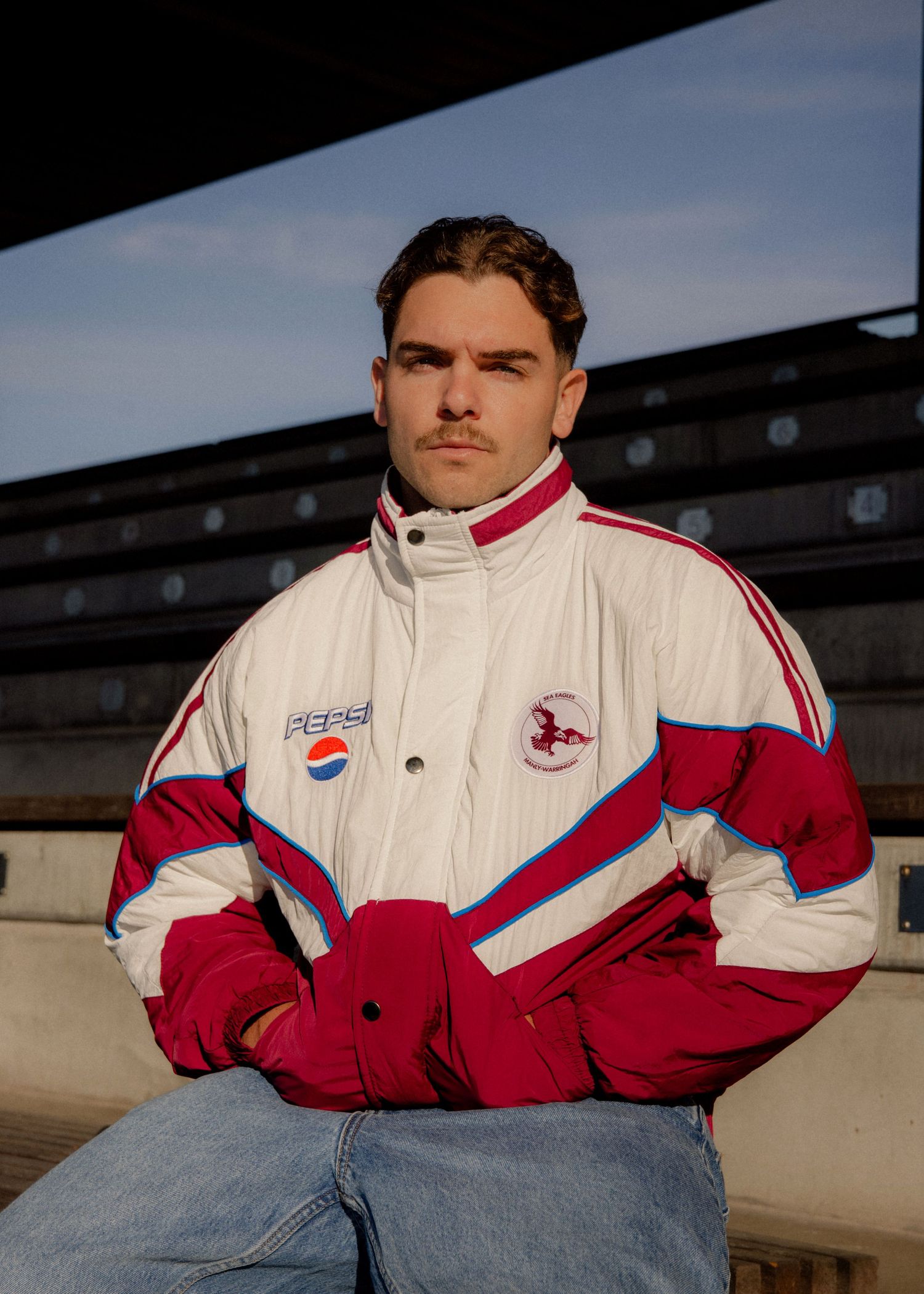 Manly Eagles NRL Jacket - Young man wearing a vintage Pepsi-branded red and white racing jacket, sitting in stadium bleachers outdoors.