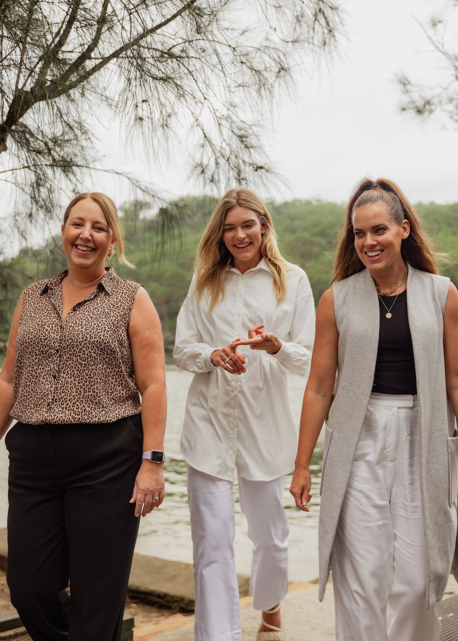 Three women walking outdoors near waterfront, smiling and dressed in casual elegant attire.