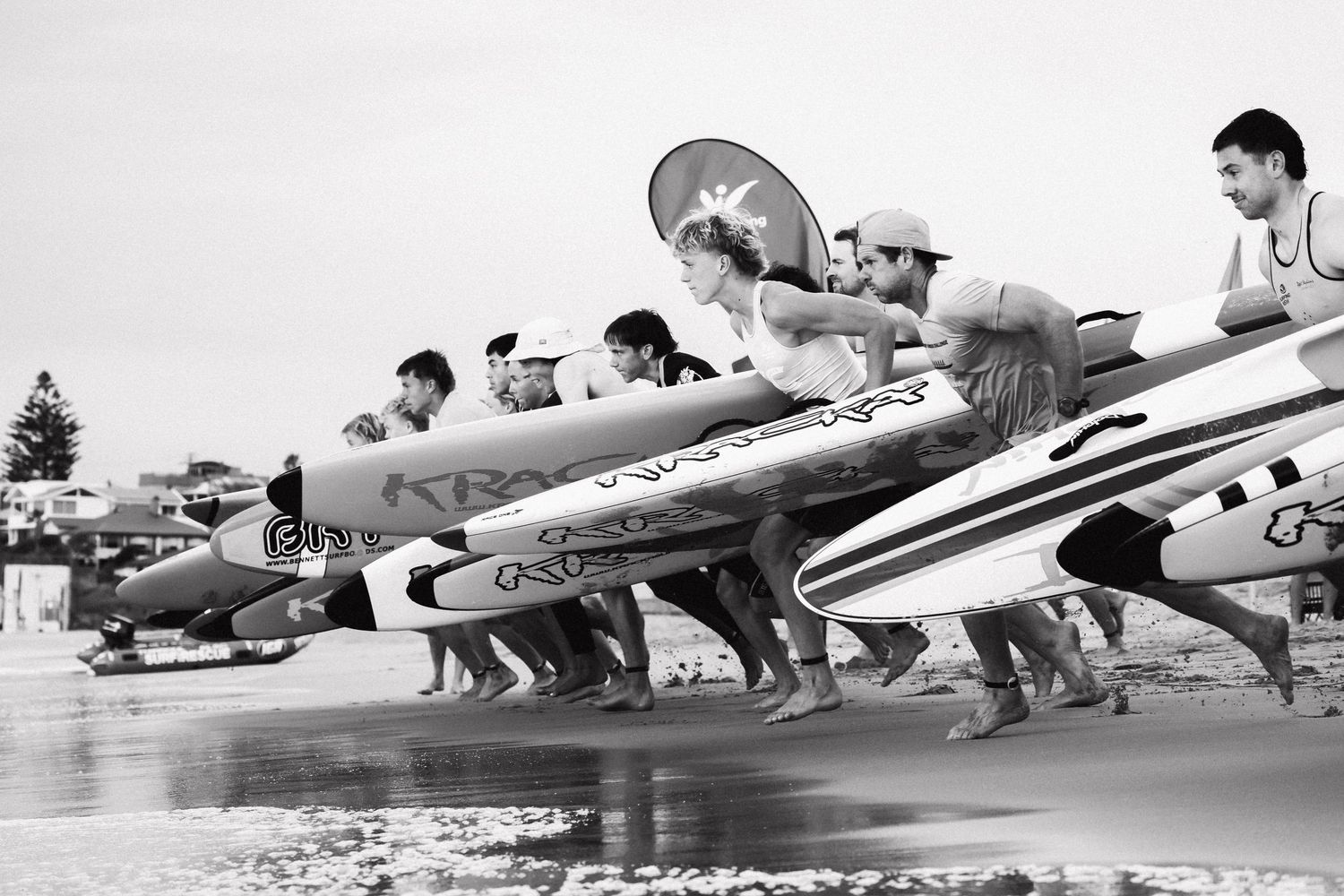 Black and white photo of surfers lined up on a beach, holding surfboards ready to enter the ocean.