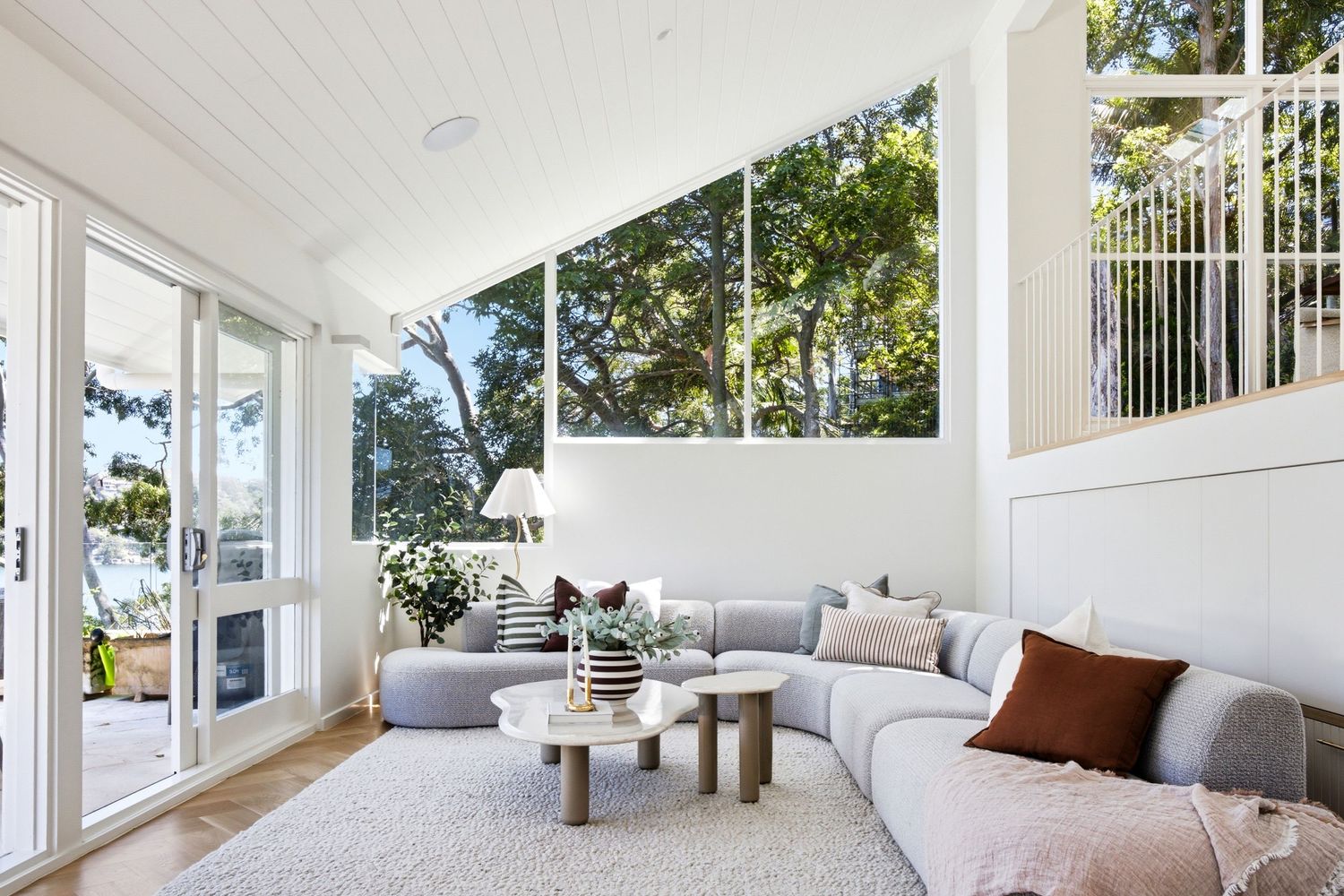 Bright sunlit living room with vaulted ceiling, large windows, grey sectional sofa, and modern coffee table overlooking lush trees.