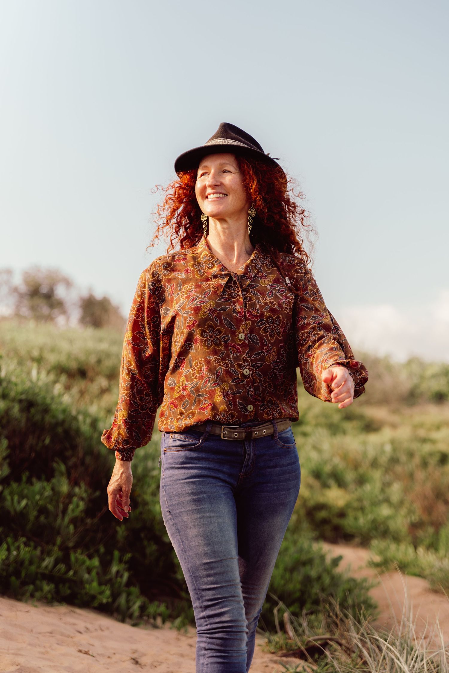 Smiling redhead woman in floral blouse, black hat, and jeans walking outdoors on a sunny day.