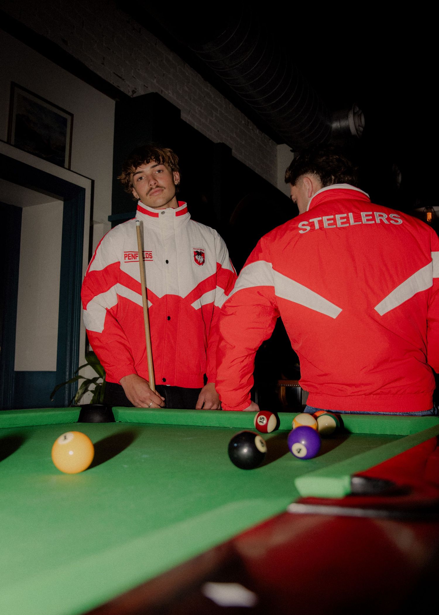 Two men in red and white Steelers NRL jackets play pool on a green billiard table. Wollongong photography