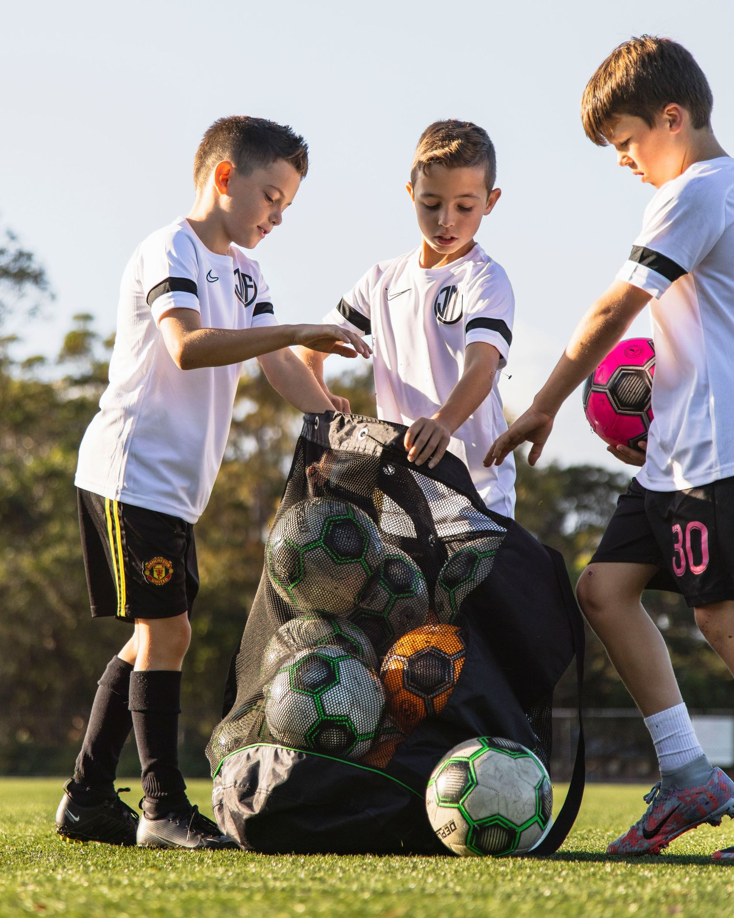 Sutherland Shire sports photographer captures Young soccer players in white uniforms gather around soccer balls on grass field.