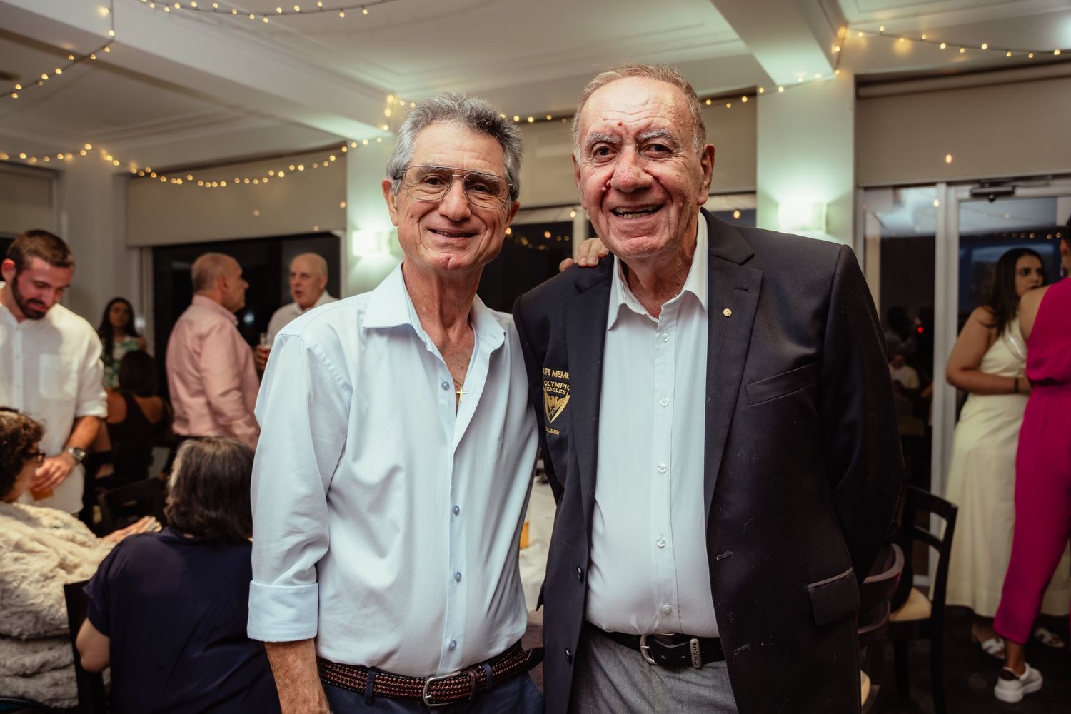 Two elderly men smiling and posing together at an indoor evening event with festive string lights in the background.