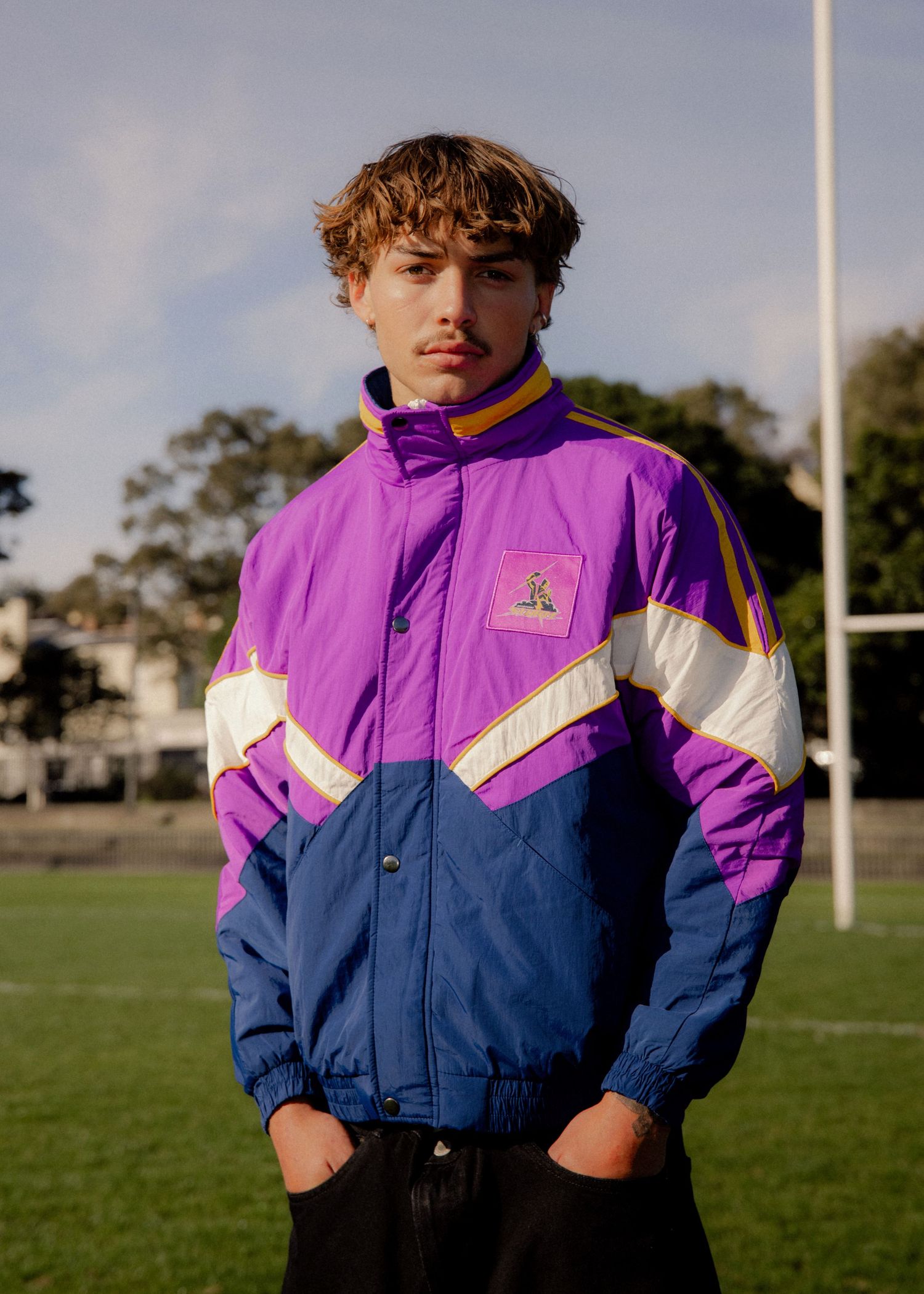 Young man standing on sports field wearing a retro purple, blue, and gold windbreaker jacket. Business and brand photographer sydney
