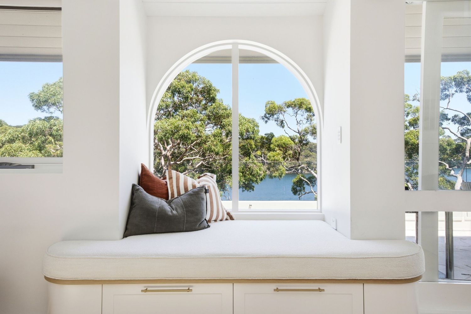 White built-in window seat with cushions and arch window overlooking ocean and eucalyptus trees.