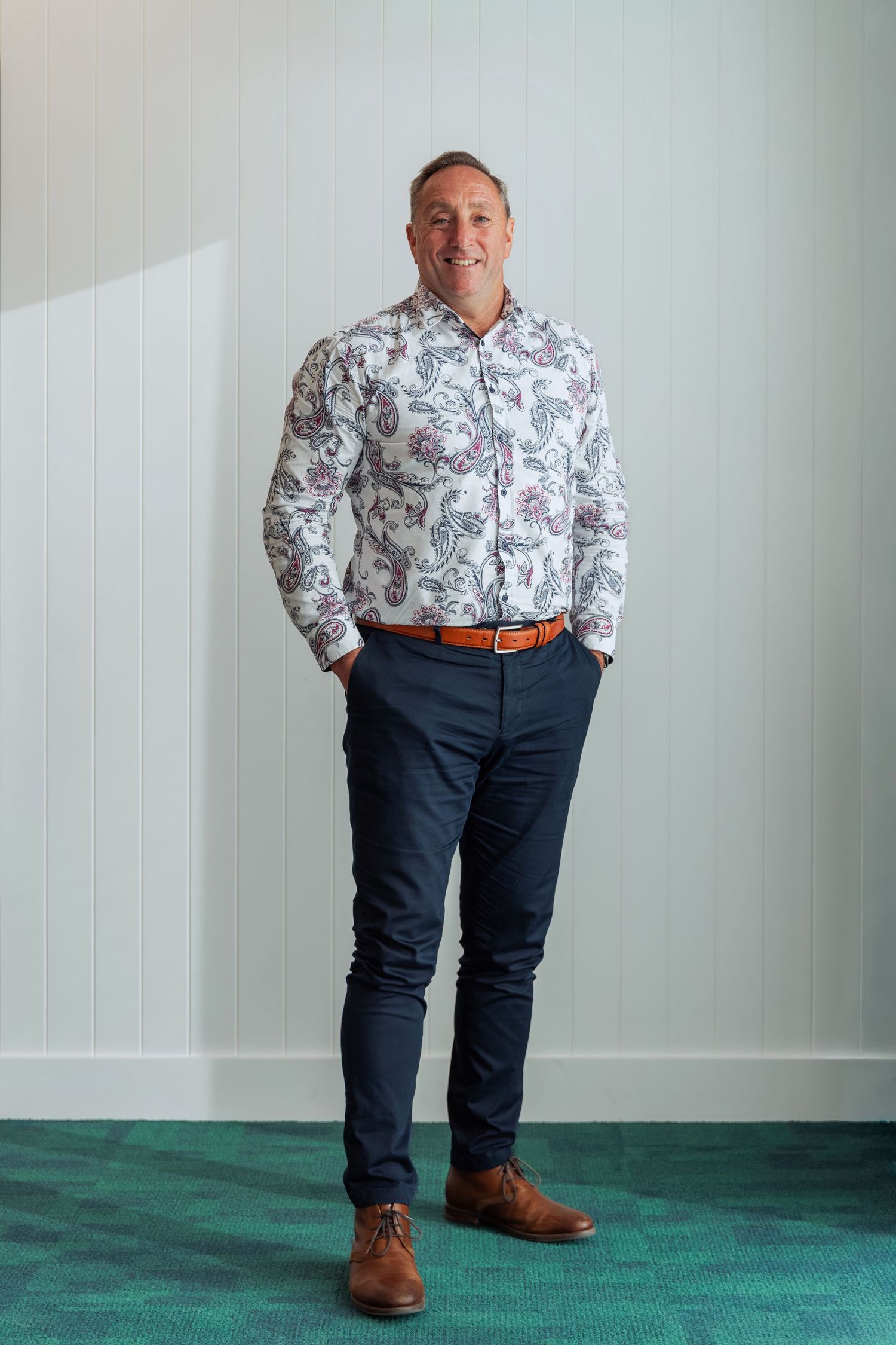 Smiling man in paisley shirt, navy chinos, and brown leather shoes standing confidently indoors.