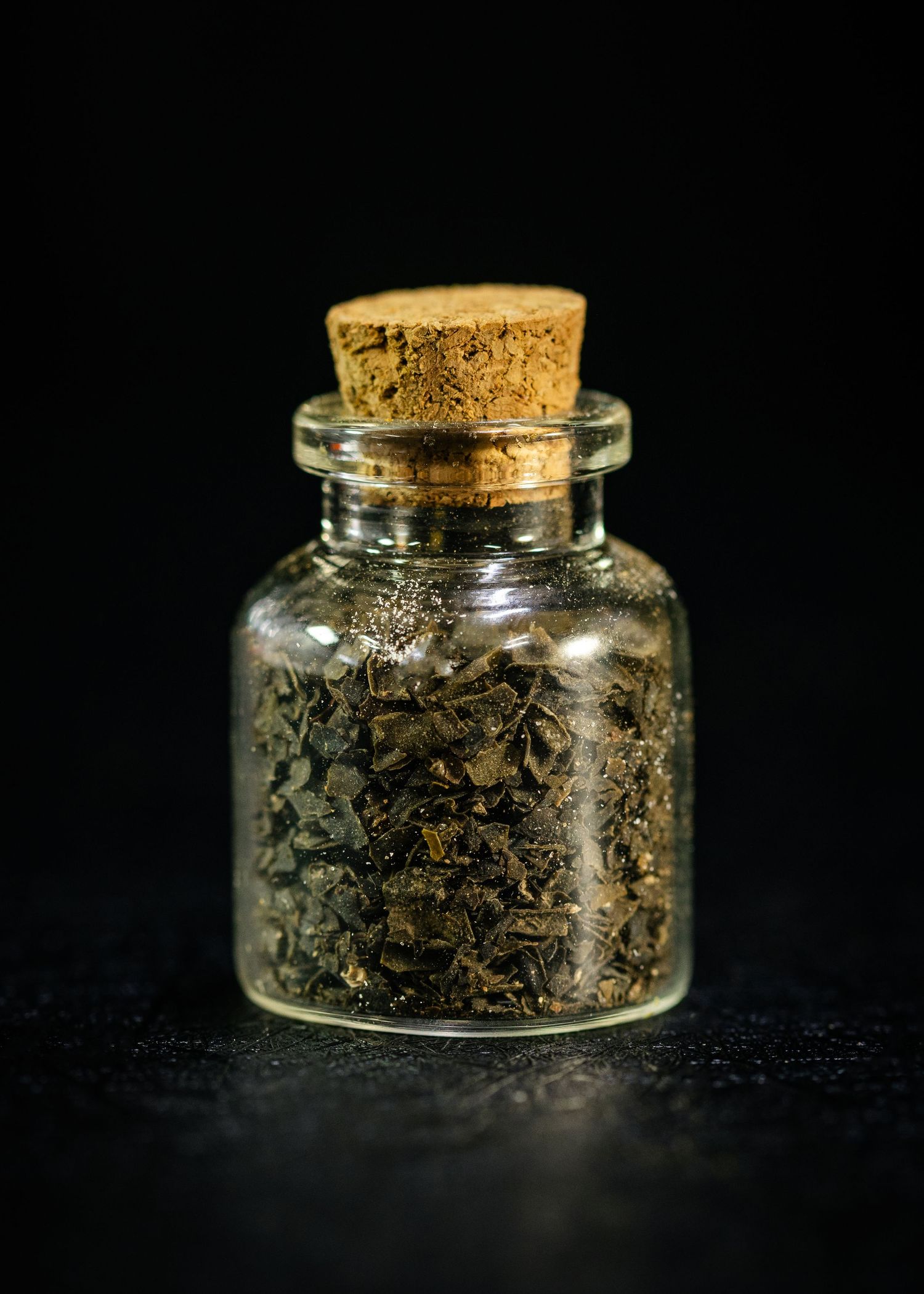 Small glass jar with cork stopper filled with dried green herbs on dark background.