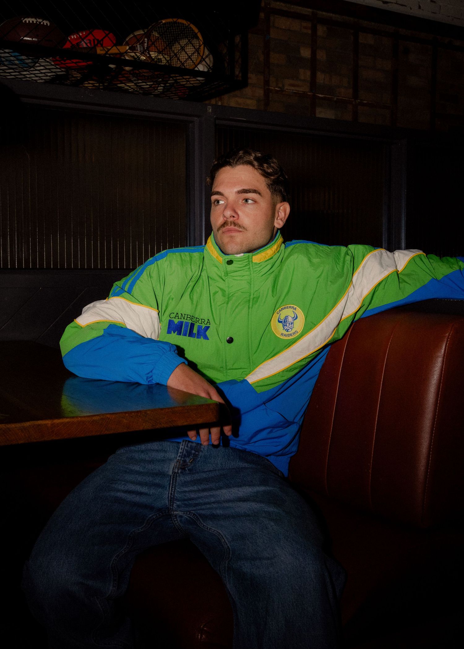 Male model wearing a retro green, white and blue Canberra Raiders NRL tracksuit jacket seated in a bar booth. Business and brand photography
