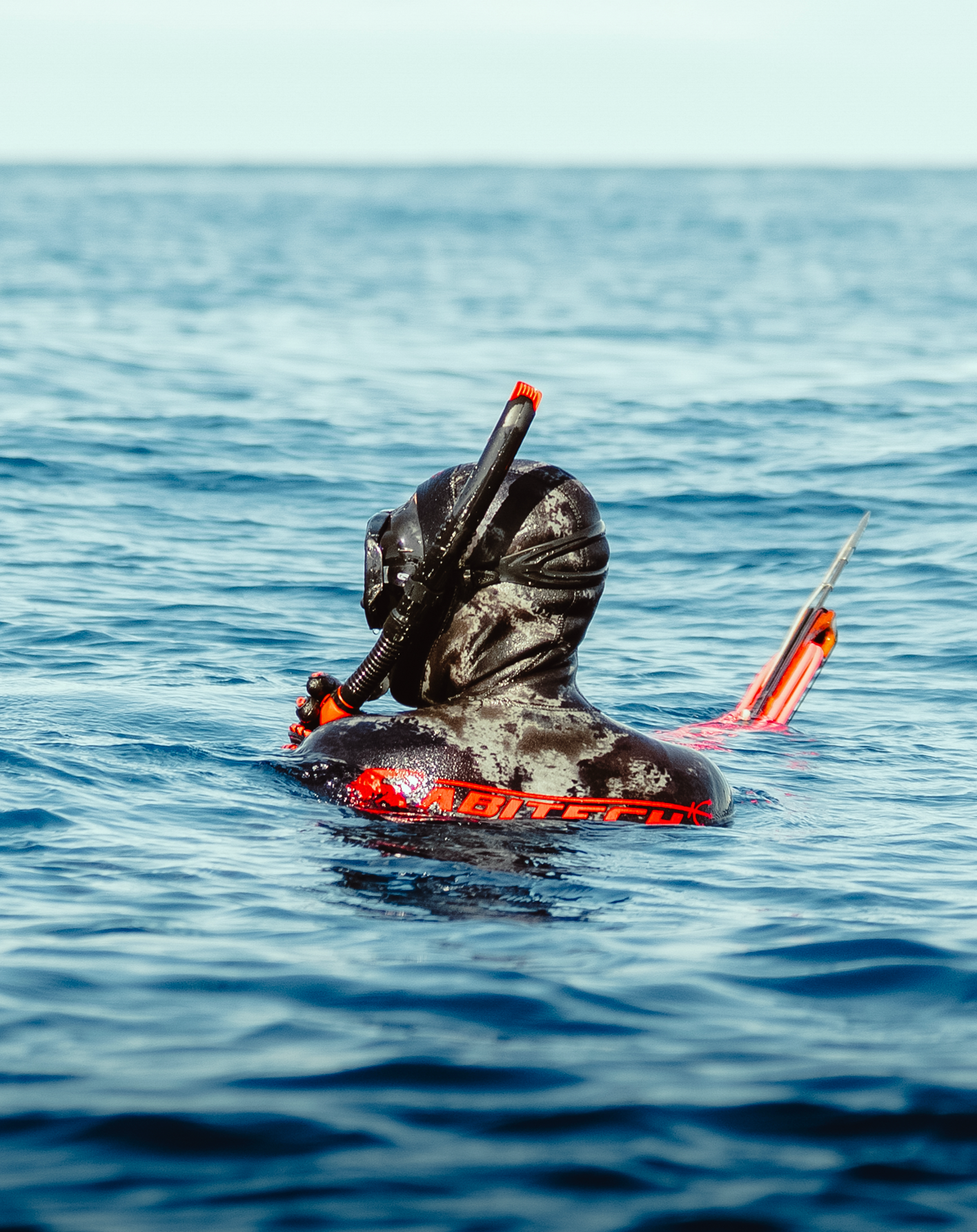 A snorkeler and spearfisherman surfaces in blue ocean water with diving gear and orange safety marker. Underwater imagery photo and video