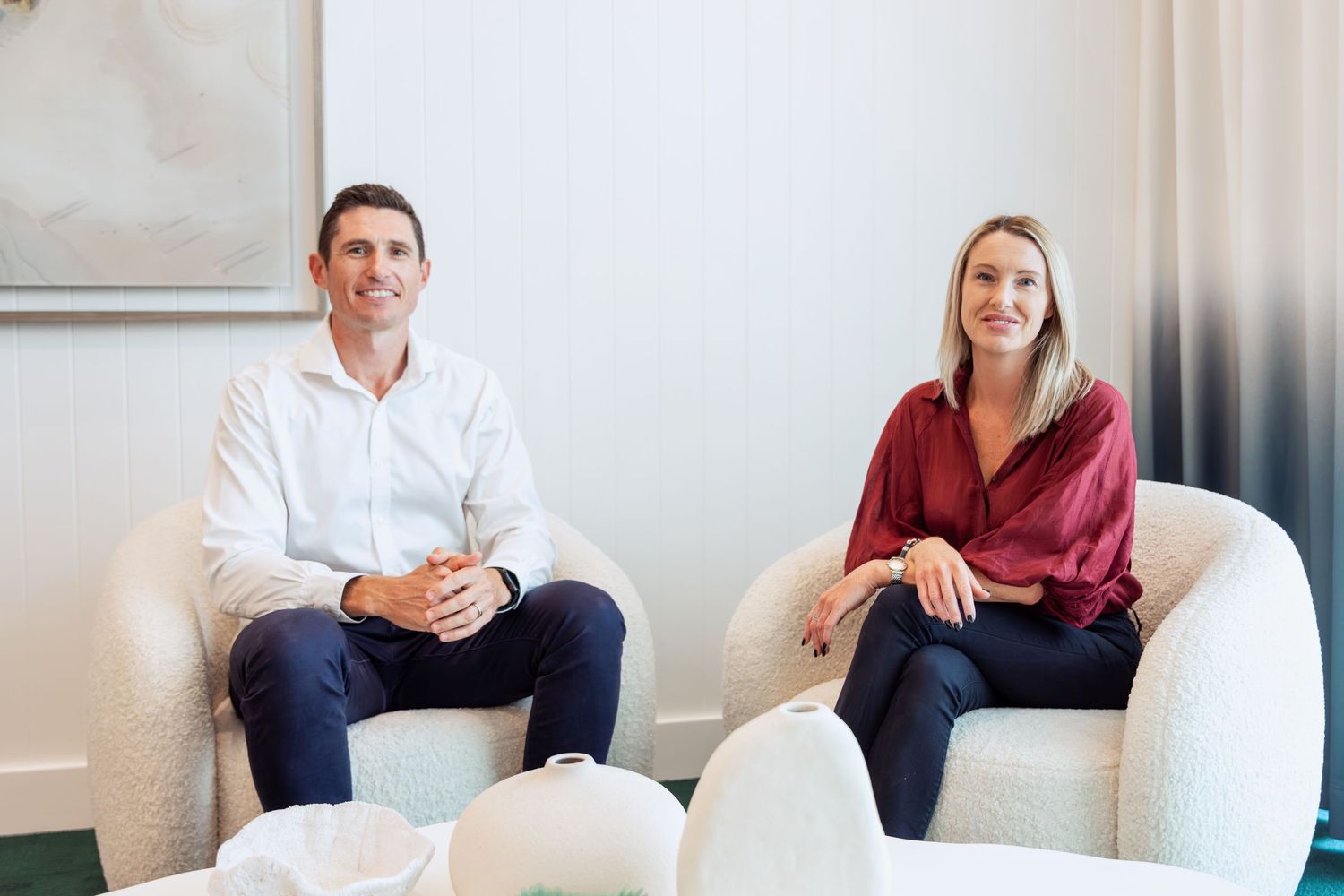 A man and woman sitting in white armchairs in a modern, bright office setting, smiling professionally.
