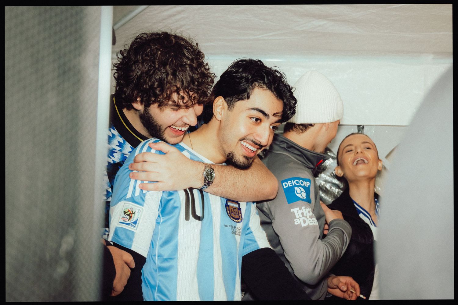 Three young people hugging and smiling in a hallway, one wearing an Argentina Adidas soccer jersey.