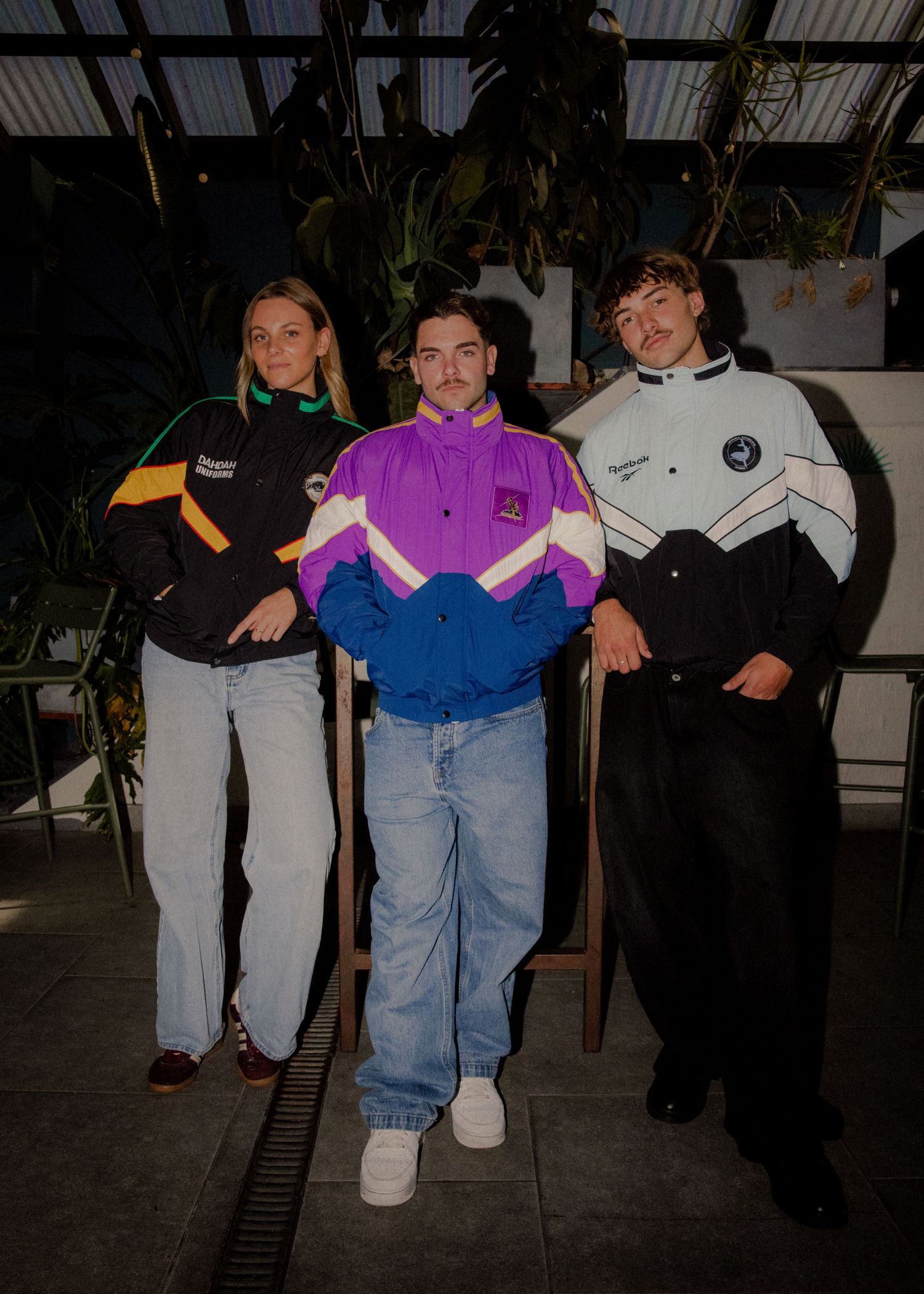 Three friends pose in colorful retro NRL windbreaker jackets inside a lush greenhouse setting. Wollongong photography