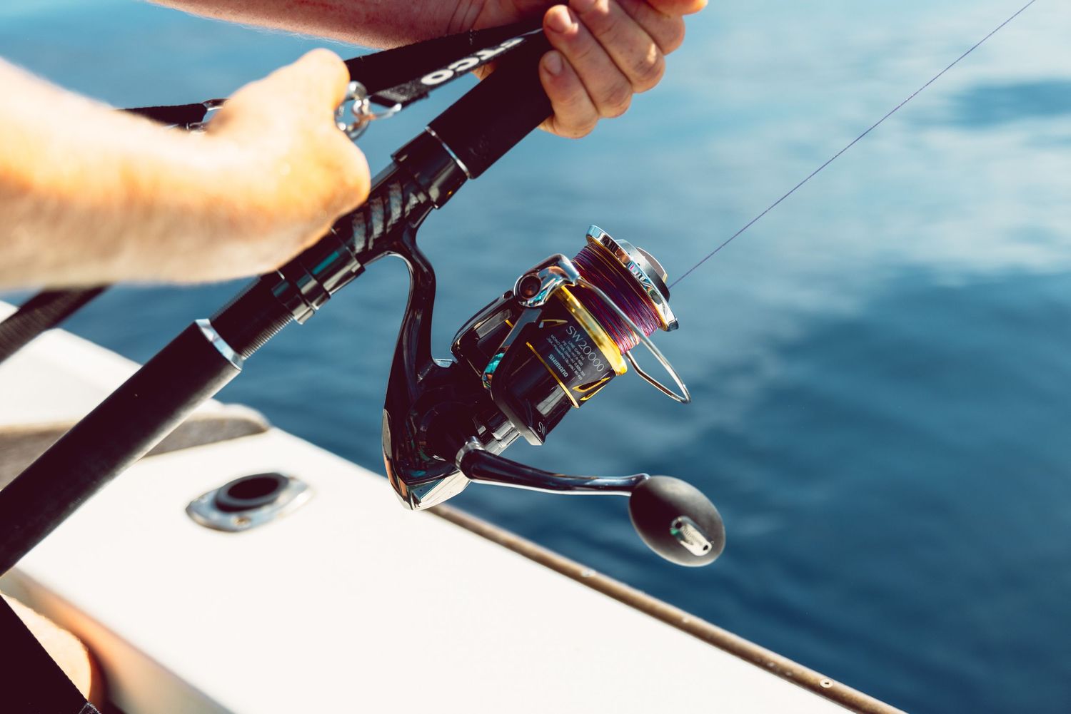 Close-up of a fishing rod and spinning reel held over calm blue ocean water from a boat deck.