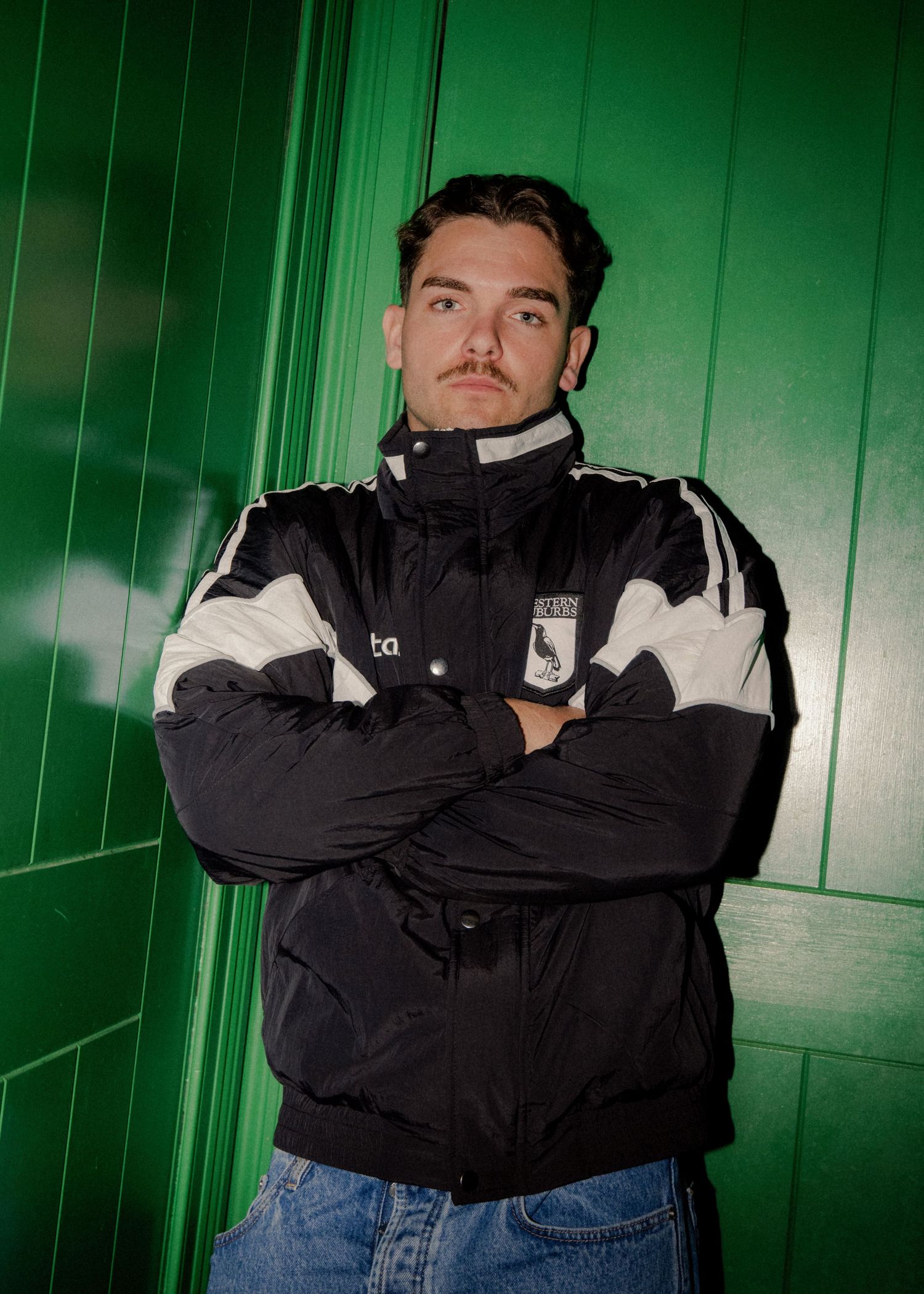 Sydney brand phototgrapher - Young man with mustache posing in black and white sports jacket with arms crossed against green wall.