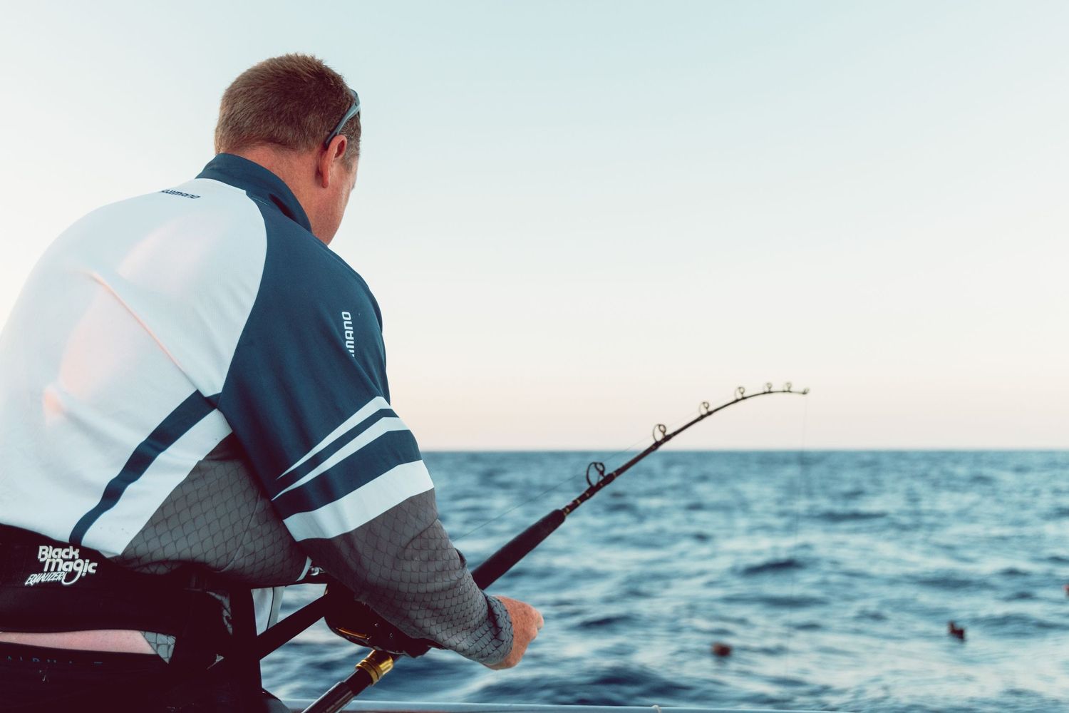 Man fishing from a boat on calm ocean waters, holding a fishing rod against a clear horizon.