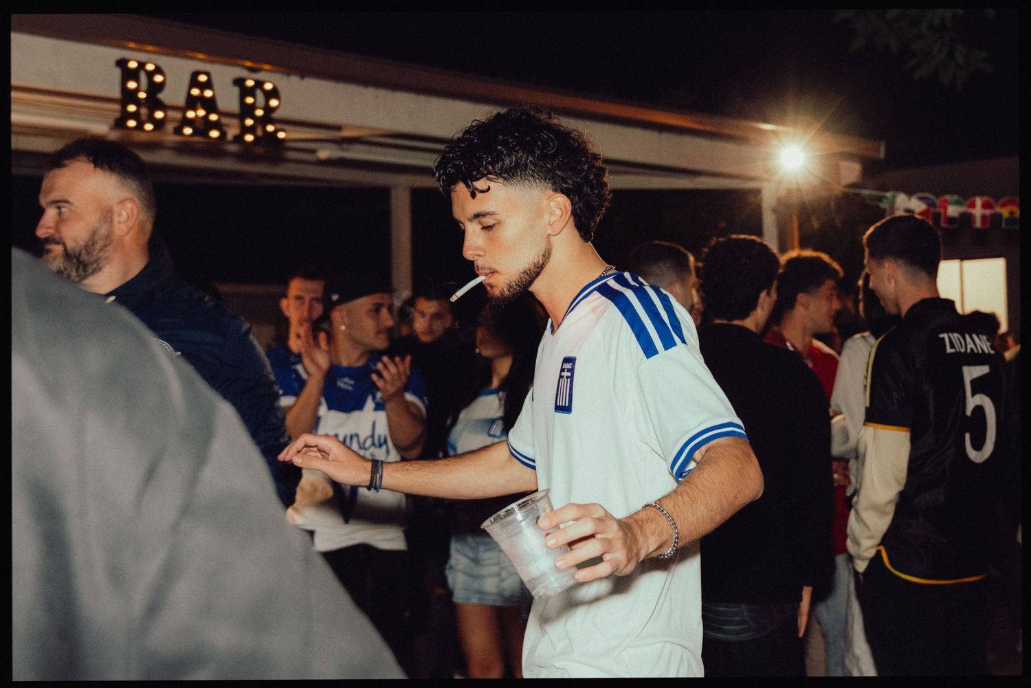 Young man in soccer jersey holding drink at crowded outdoor bar party at night.