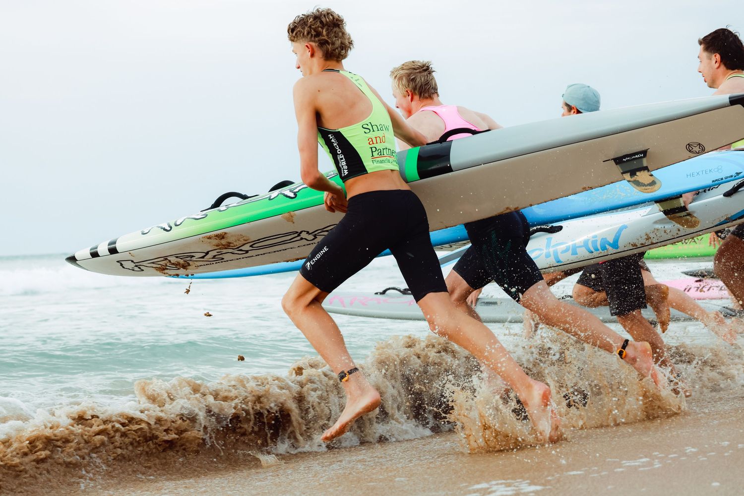Young surfers running into ocean waves carrying colorful surfboards during a beach competition on a sunny day.