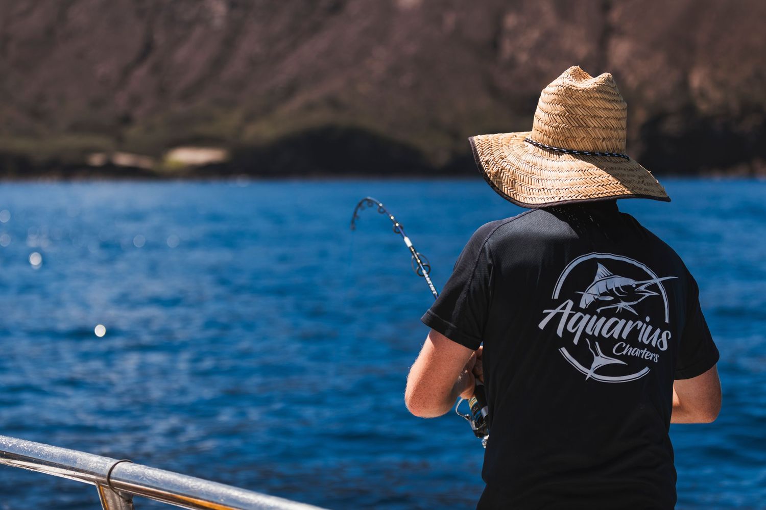 Person wearing Aquarius Cruises black t-shirt and straw hat fishing from a boat with scenic ocean views.