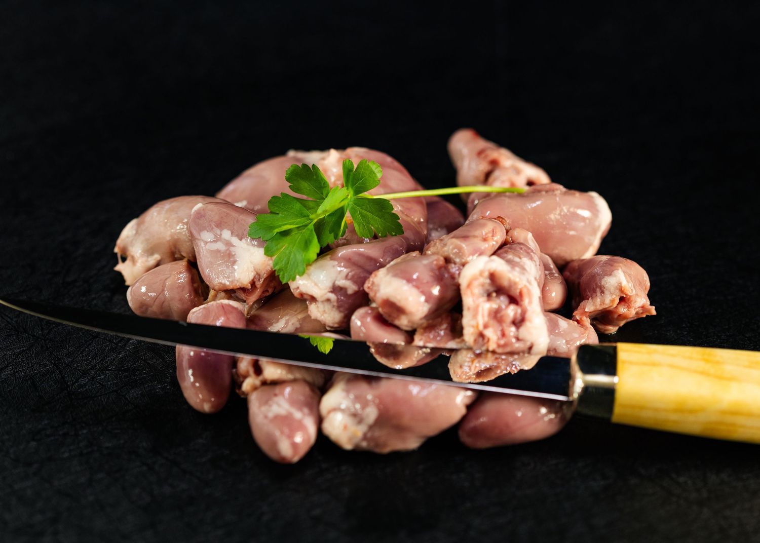 Raw chicken hearts piled on a knife blade, garnished with fresh parsley, on a dark background.
