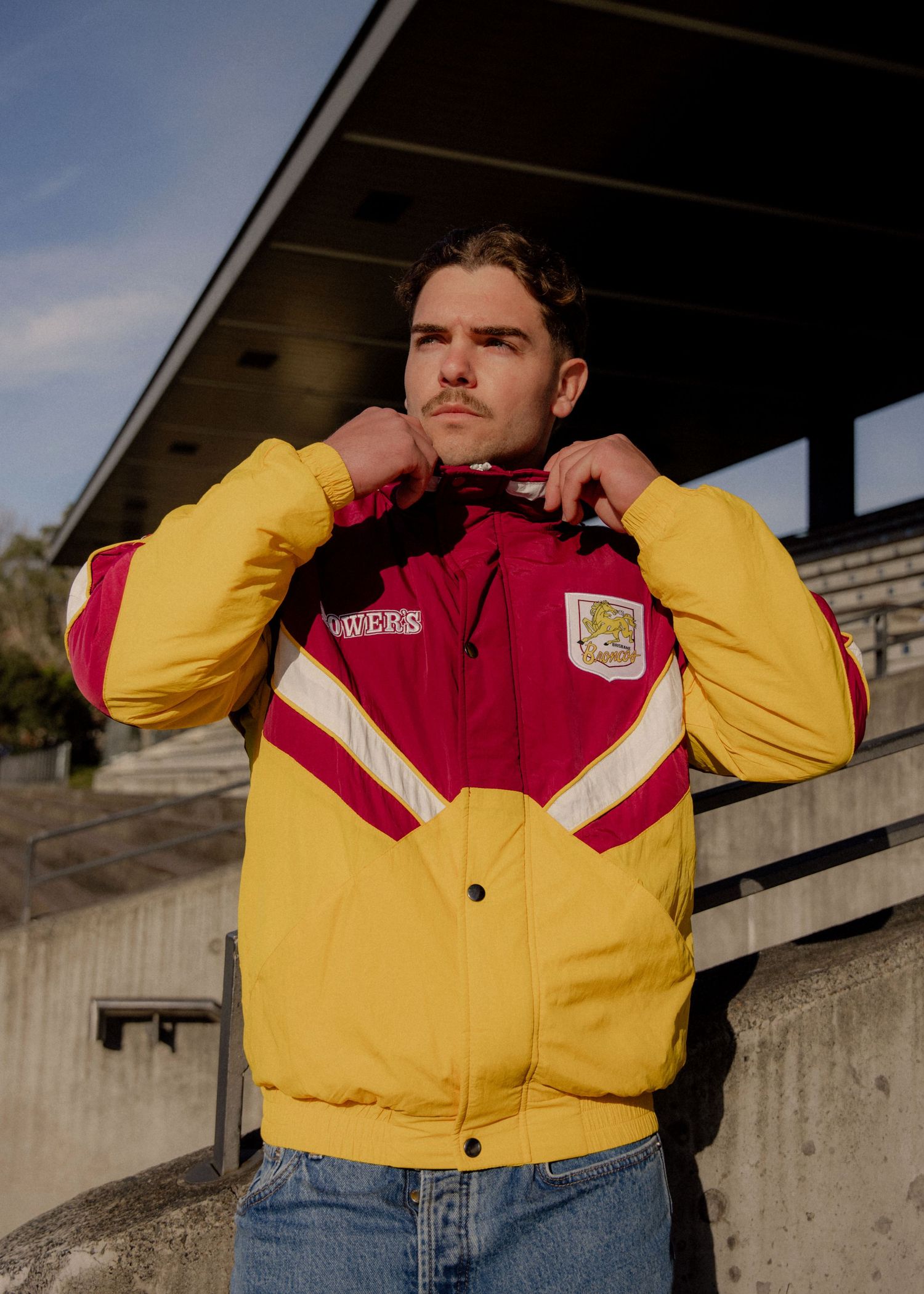 Man wearing yellow and red retro sports jacket adjusting collar outdoors near stadium bleachers.