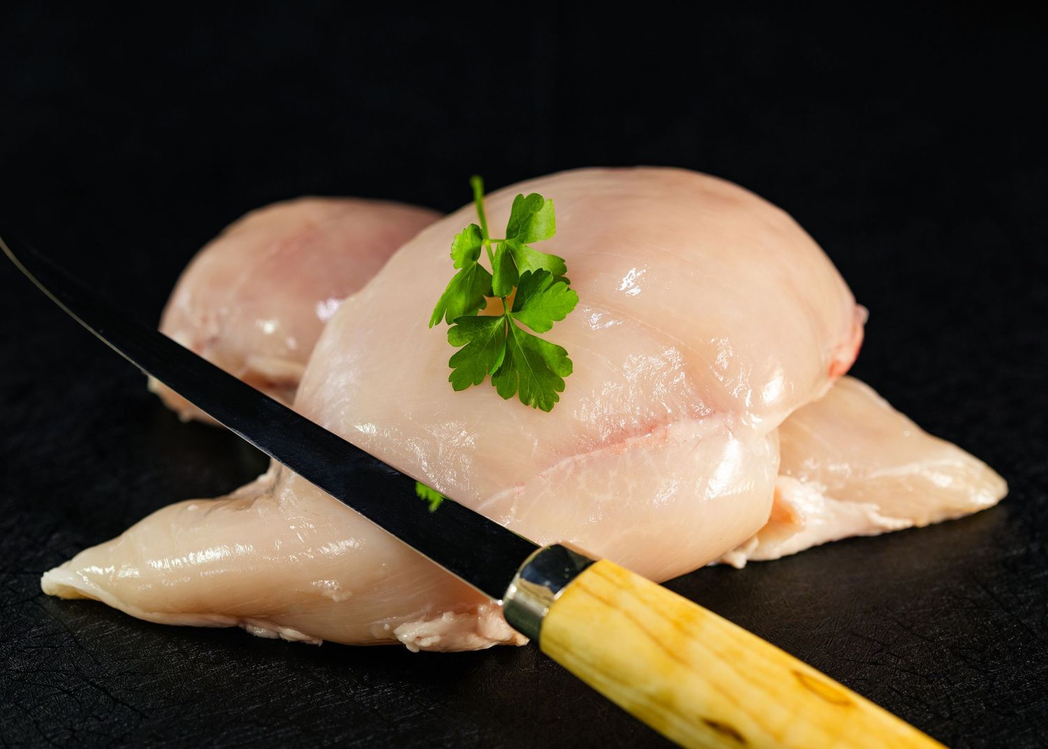 Food and product photography and videography Fresh raw chicken breasts garnished with parsley and a wooden-handled knife on black surface.