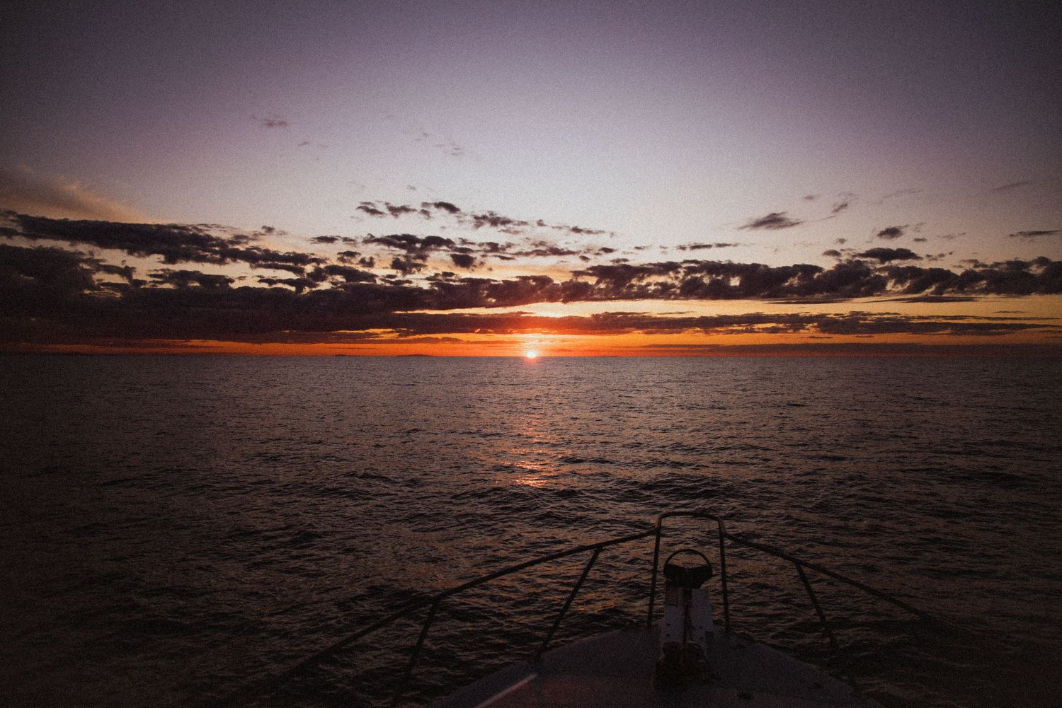 A stunning ocean sunset viewed from a boat bow, with vibrant orange horizon and dramatic clouds over calm waters.