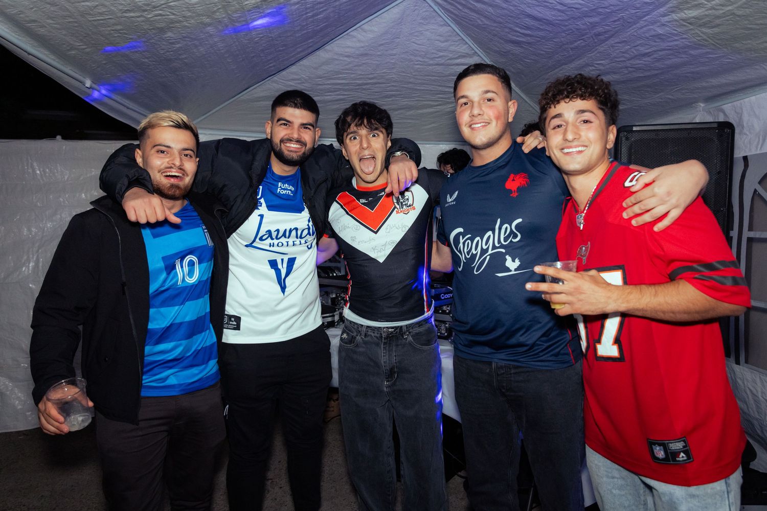 Five young men pose together smiling at a party, wearing sports jerseys in a white tent venue with blue lighting.