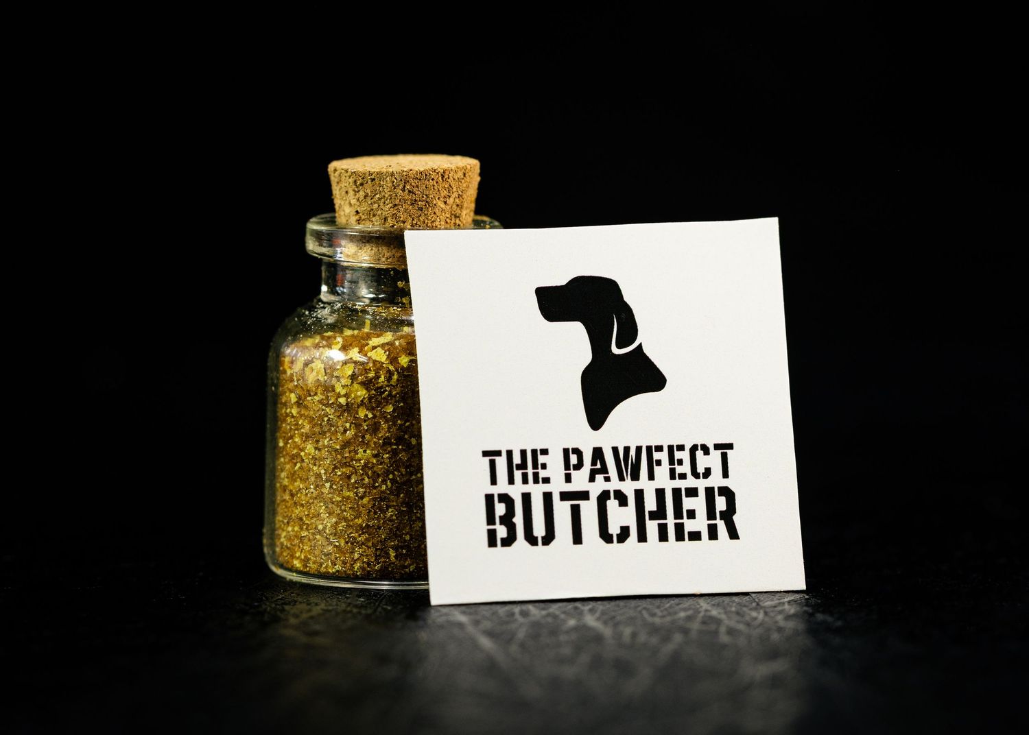 The Pawfect Butcher logo card beside a small glass jar filled with golden spices on a dark background.