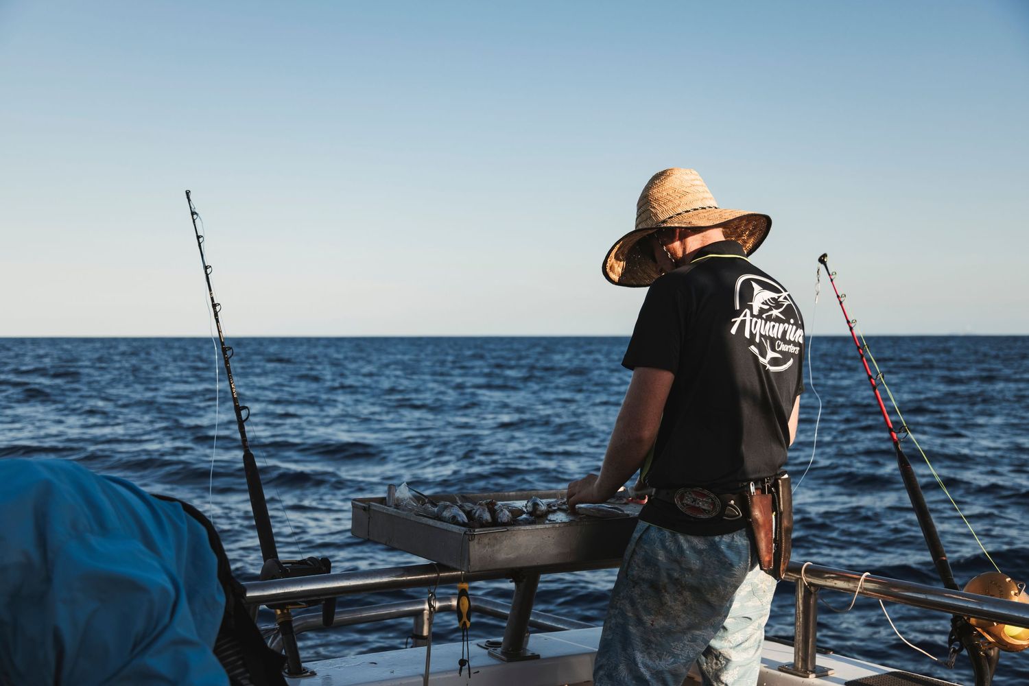 A fisherman in a straw hat stands on a boat deck surrounded by fishing rods with the open ocean in the background.