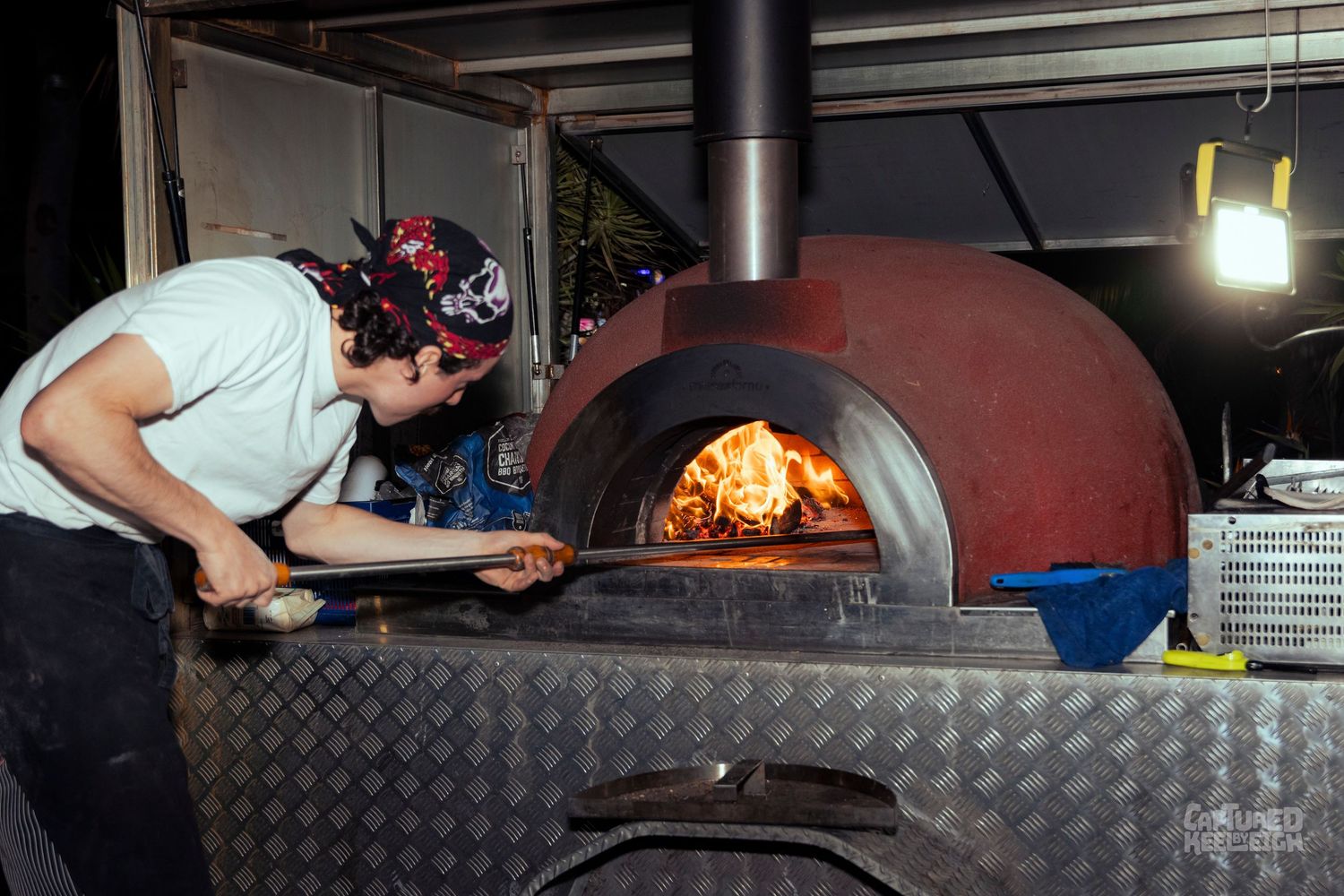 Chef using a wooden peel to tend a pizza in a traditional wood-fired oven with open flames.
