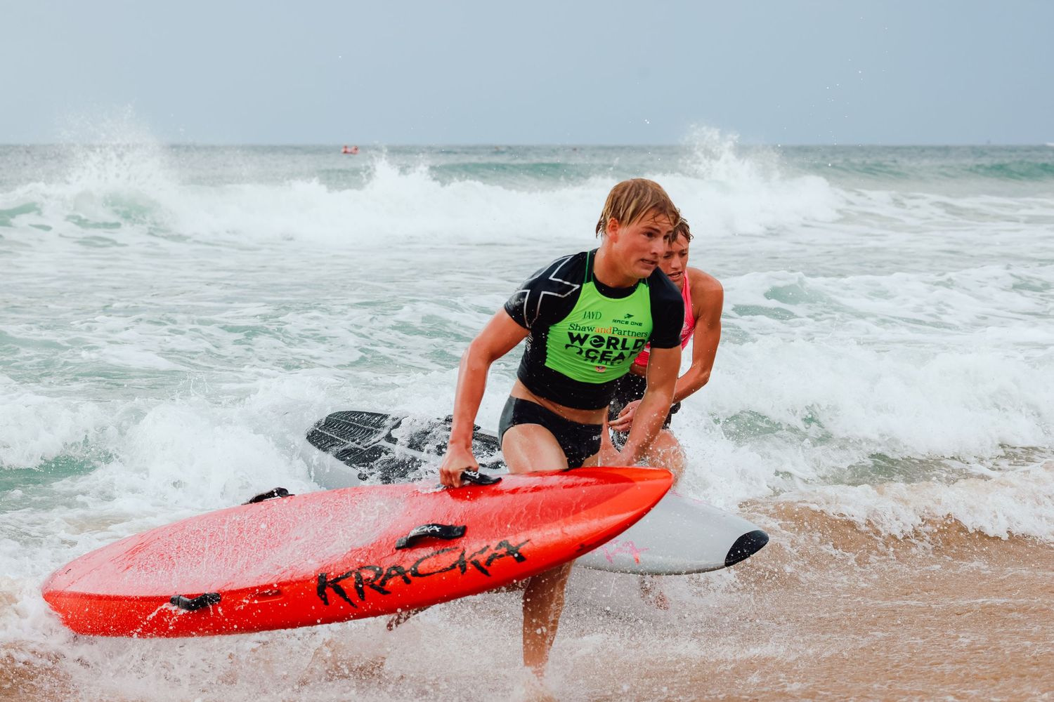 Young surfer carrying a red Kracker surfboard out of ocean waves during a world surf competition.