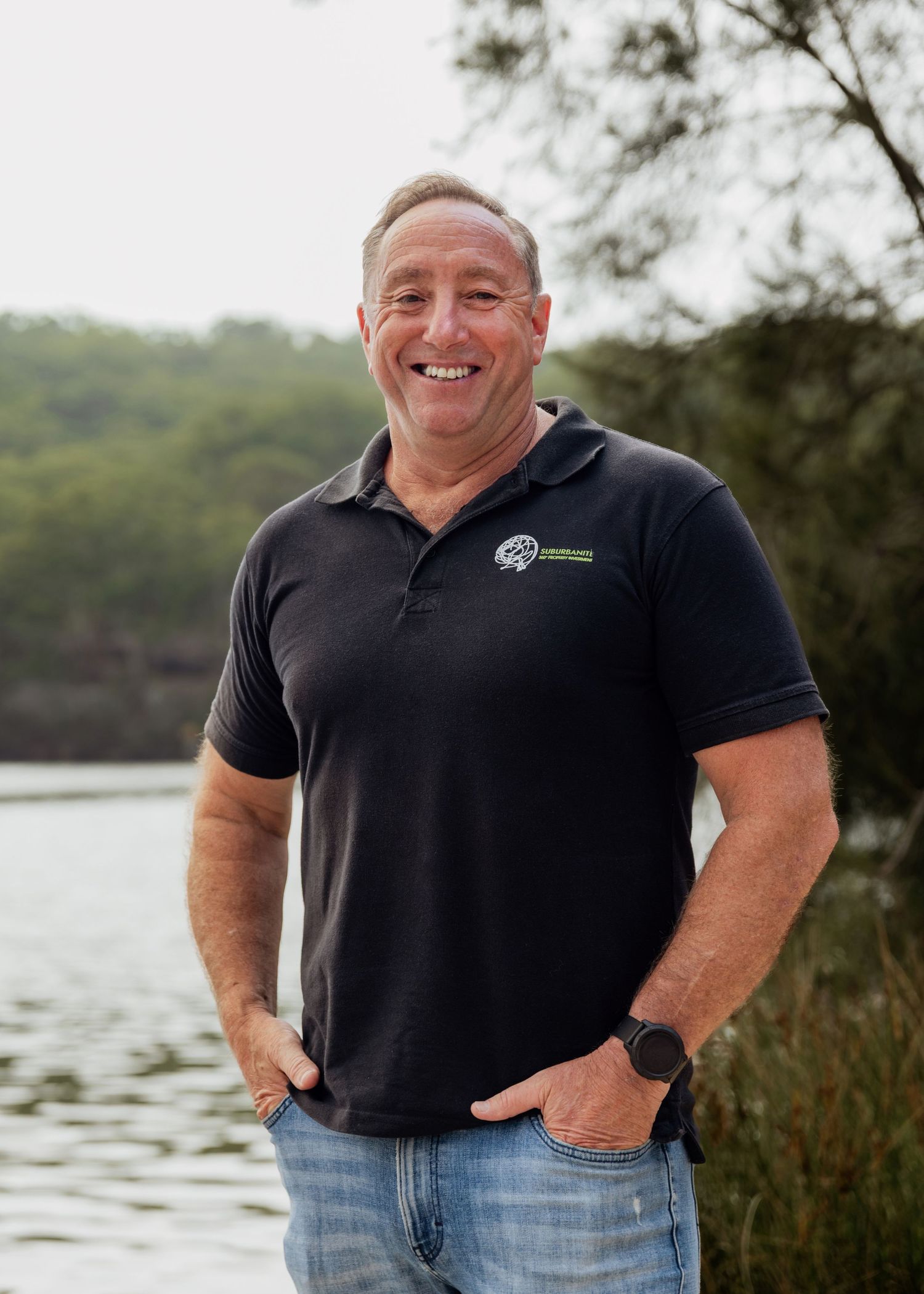 Man in navy polo shirt smiling outdoors near a lake with lush green trees in the background.