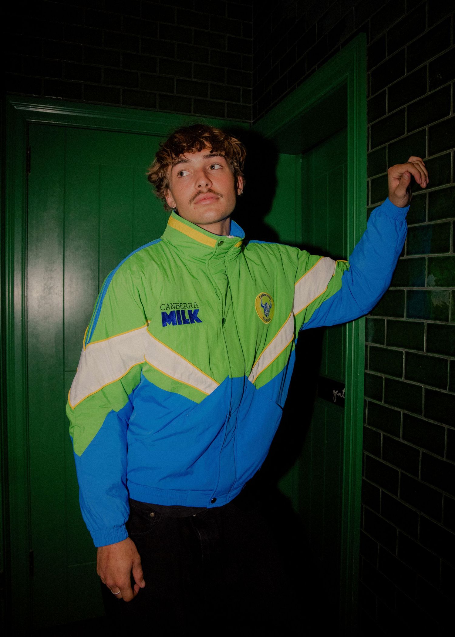 Man posing in retro green and blue tracksuit jacket with Farmer's Milk logo near green door. Sutherland Shire photographer