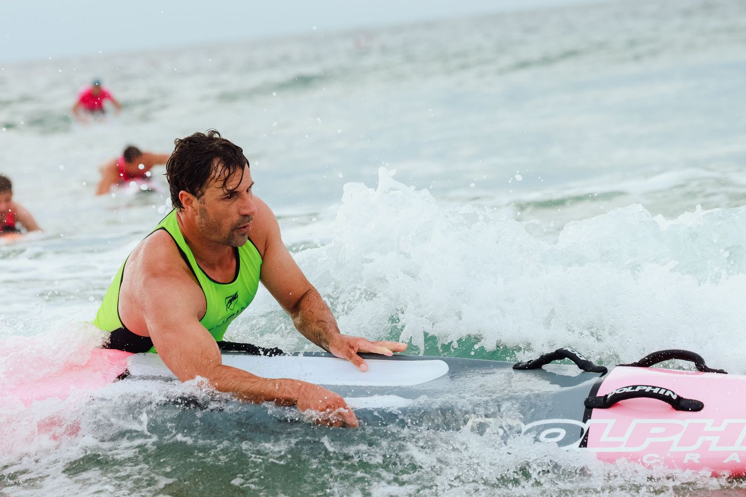 Man in green vest paddling a surfboard through ocean waves during a surfing competition.