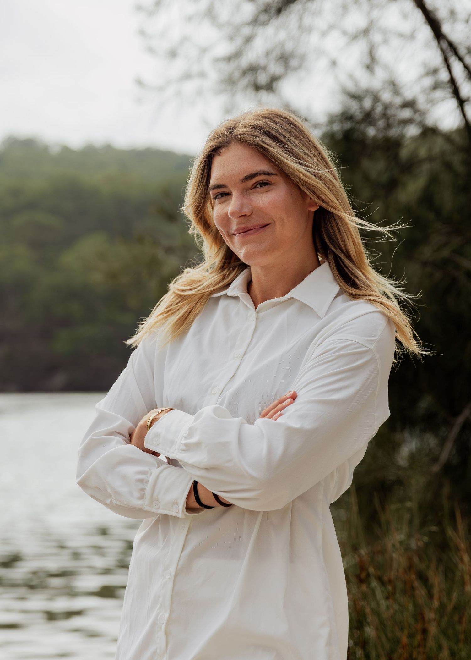 Woman in white shirt standing arms crossed by a serene lake with lush green trees in background.